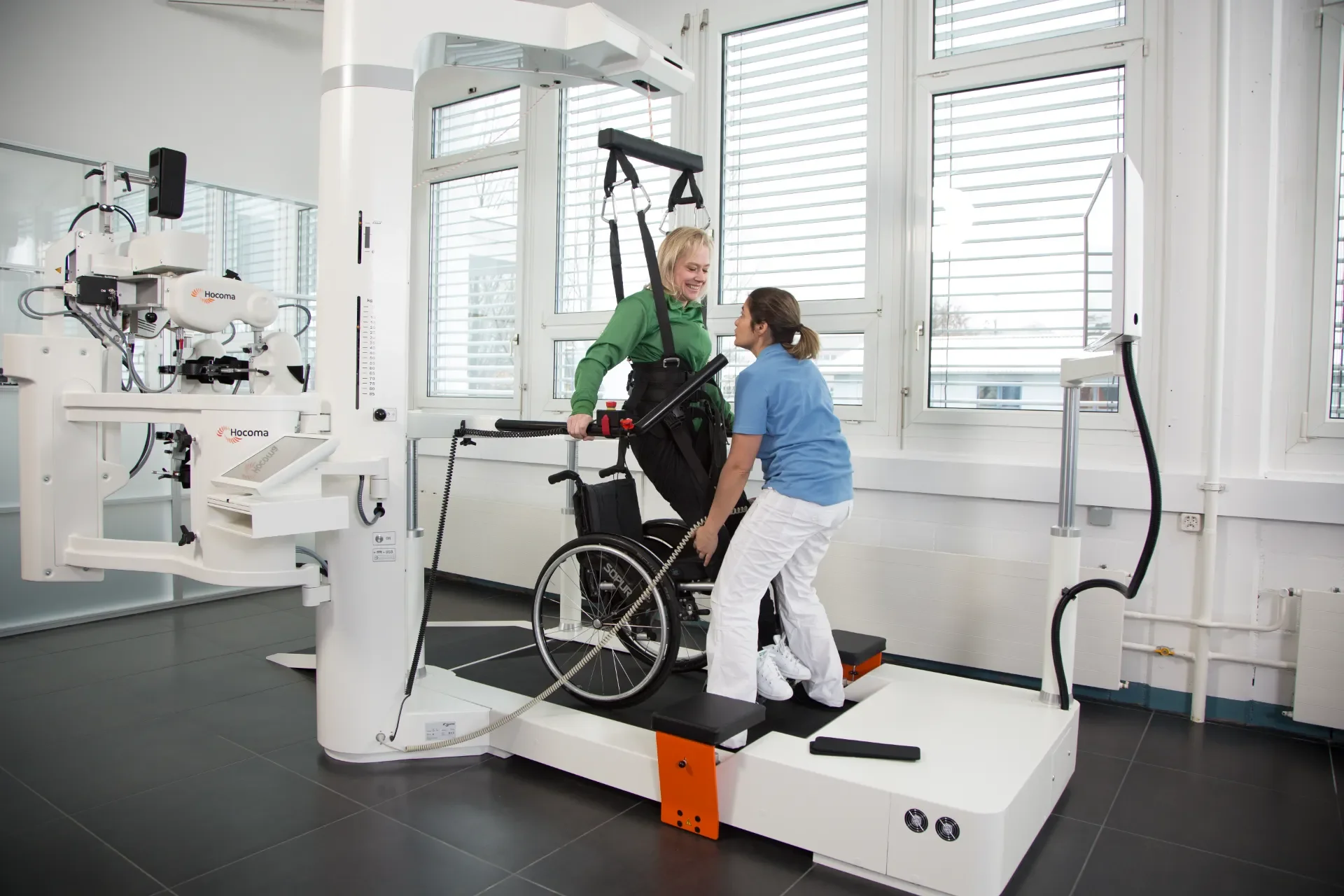 Fisioterapeuta asistiendo la transferencia de un paciente en silla de ruedas al sistema Lokomat Pro para comenzar la marcha asistida por robot. Distribuido en España y Portugal por Bayber