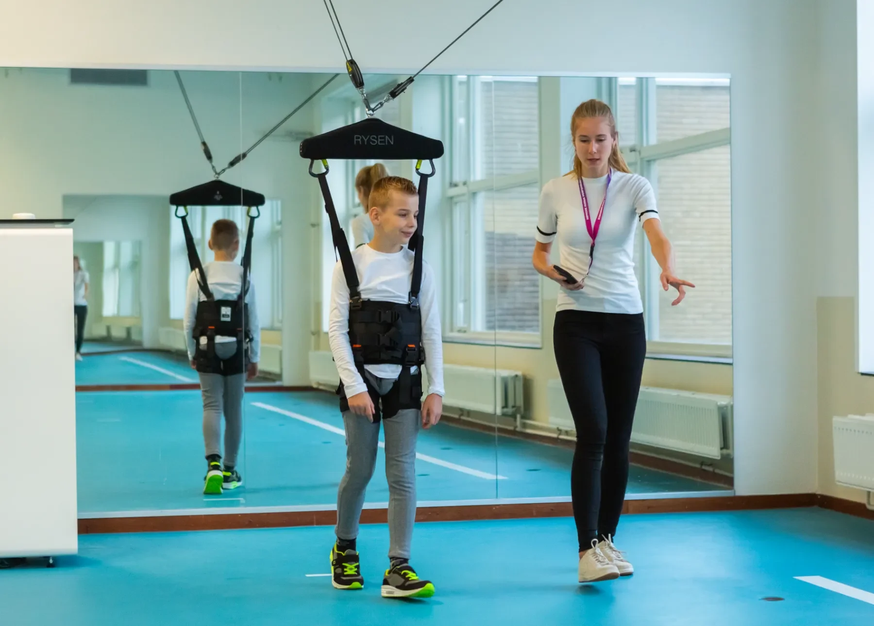 Niño caminando con soporte de RYSEN frente a espejo durante neurorrehabilitación avanzada de la marcha junto a la terapeuta. Bayber distribuidor oficial en España y Portugal