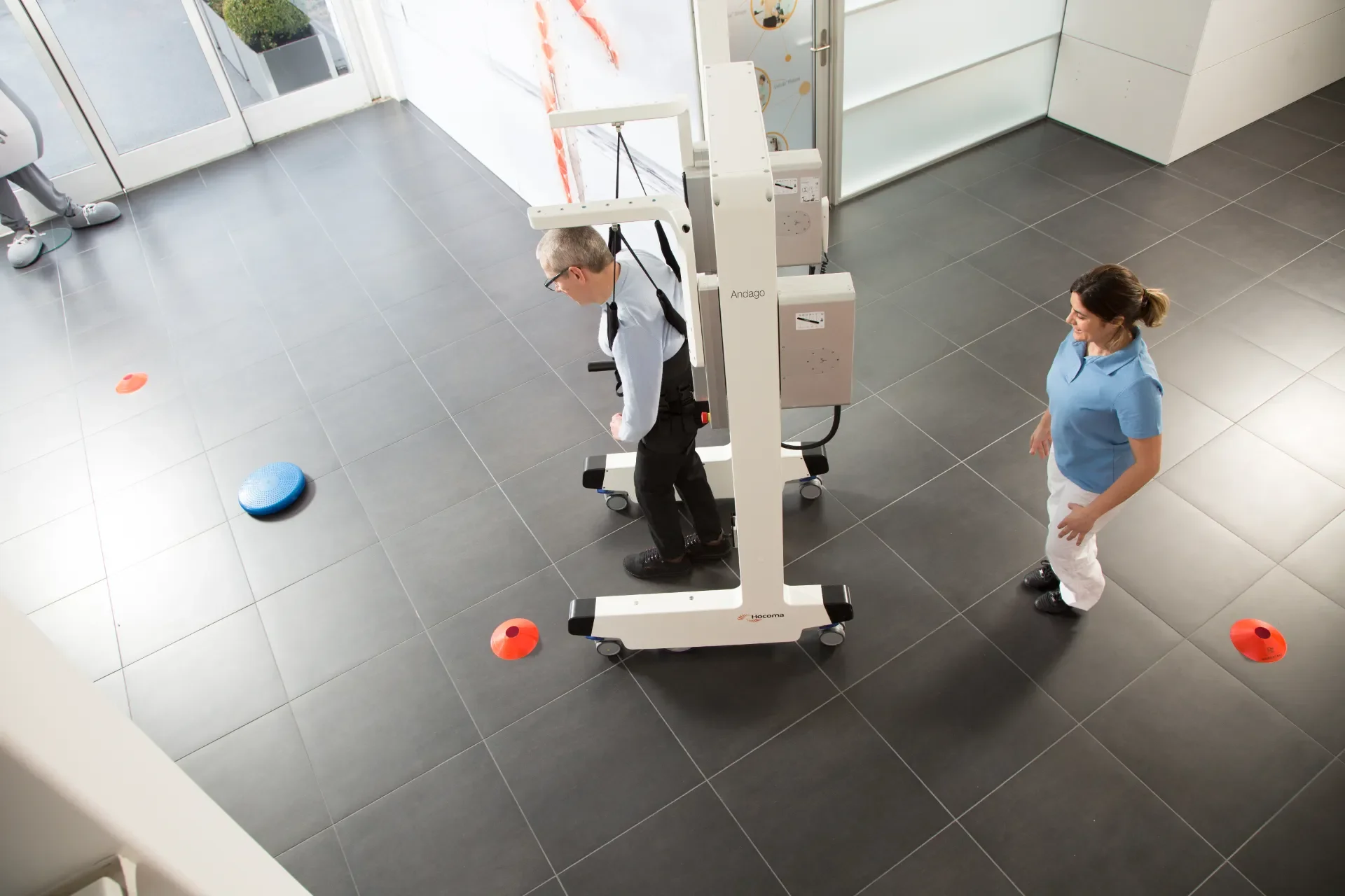 Vista cenital de paciente con lesión medular caminando con Andago 2.0 mientras la fisioterapeuta supervisa la marcha y la prevención de caídas. Bayber distribuye Andago en España y Portugal