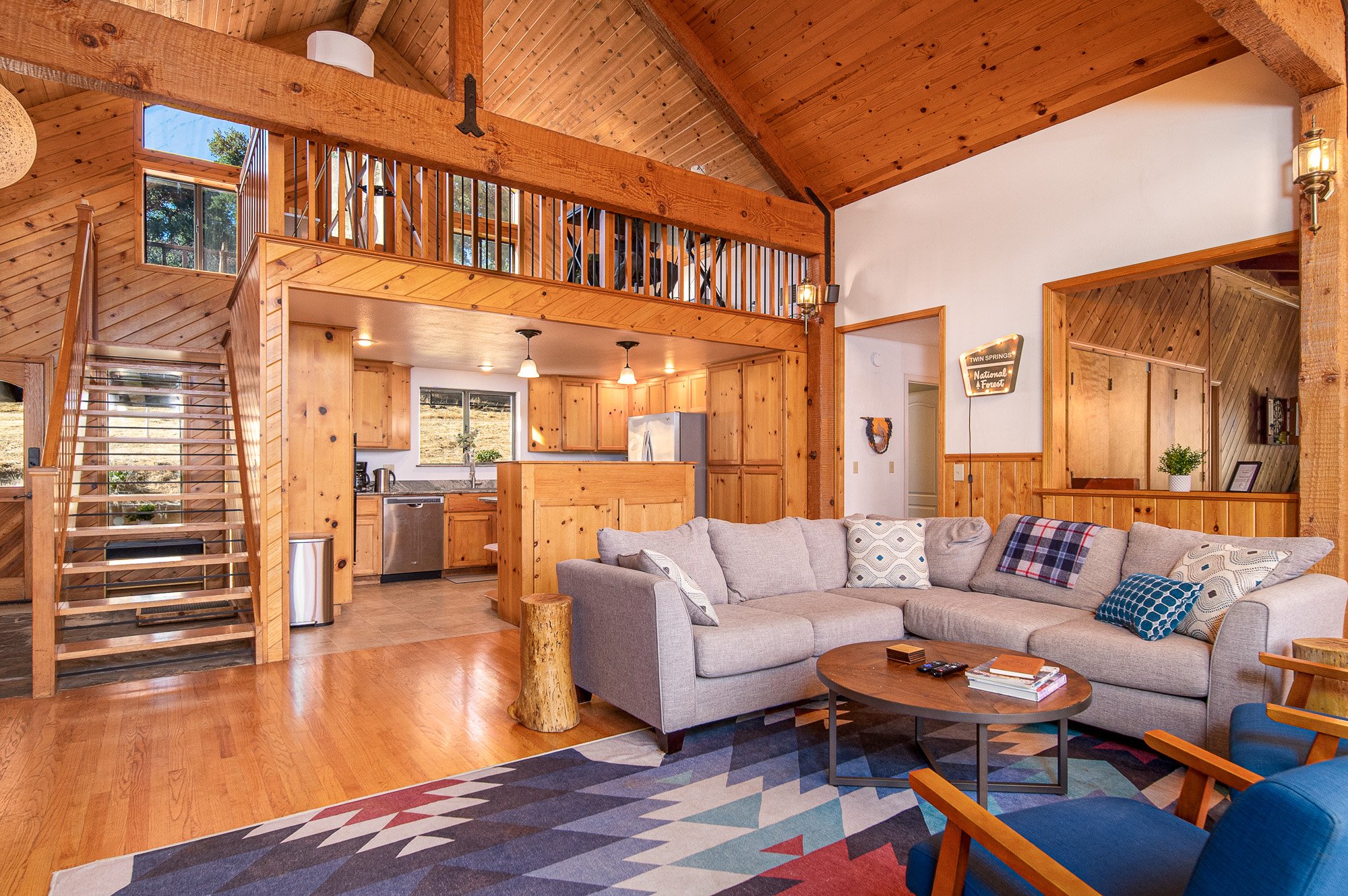 Cozy cabin interior with wooden walls, a loft, and open living space featuring a gray sectional sofa, colorful rug, and kitchen area with light wood cabinetry.