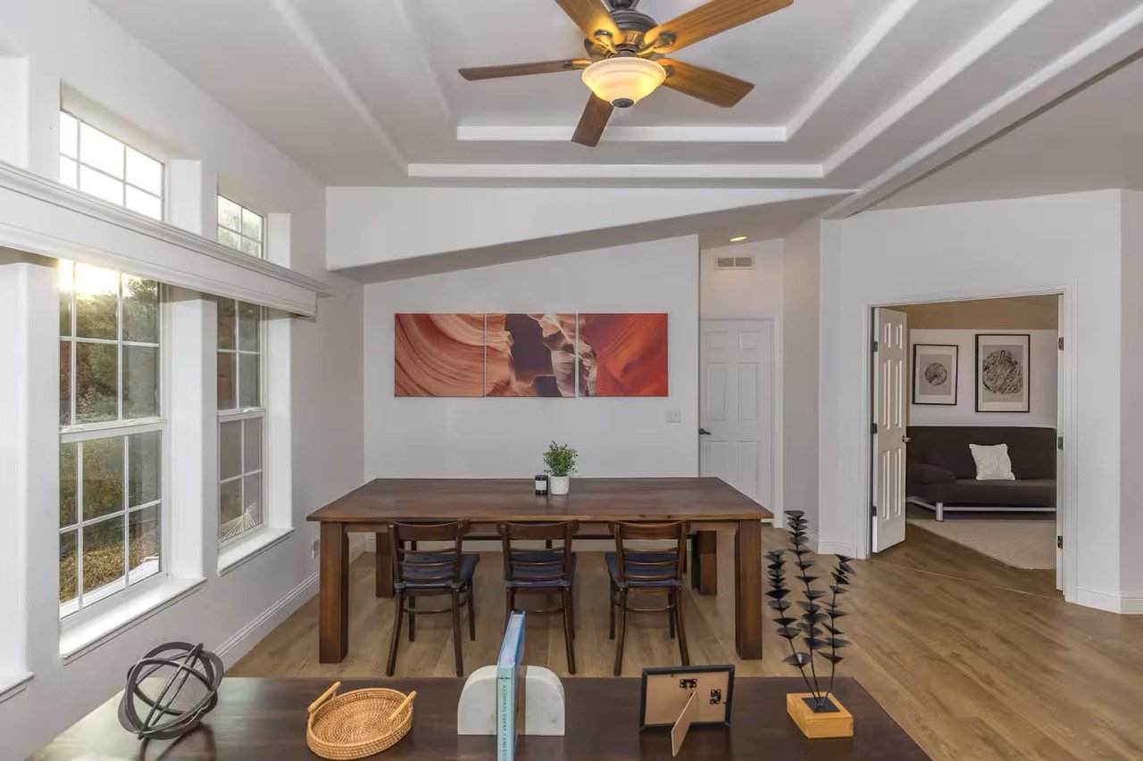 Dining room with dark wooden table and chairs, ceiling fan, large windows, abstract wall art, and adjacent living area with couch.