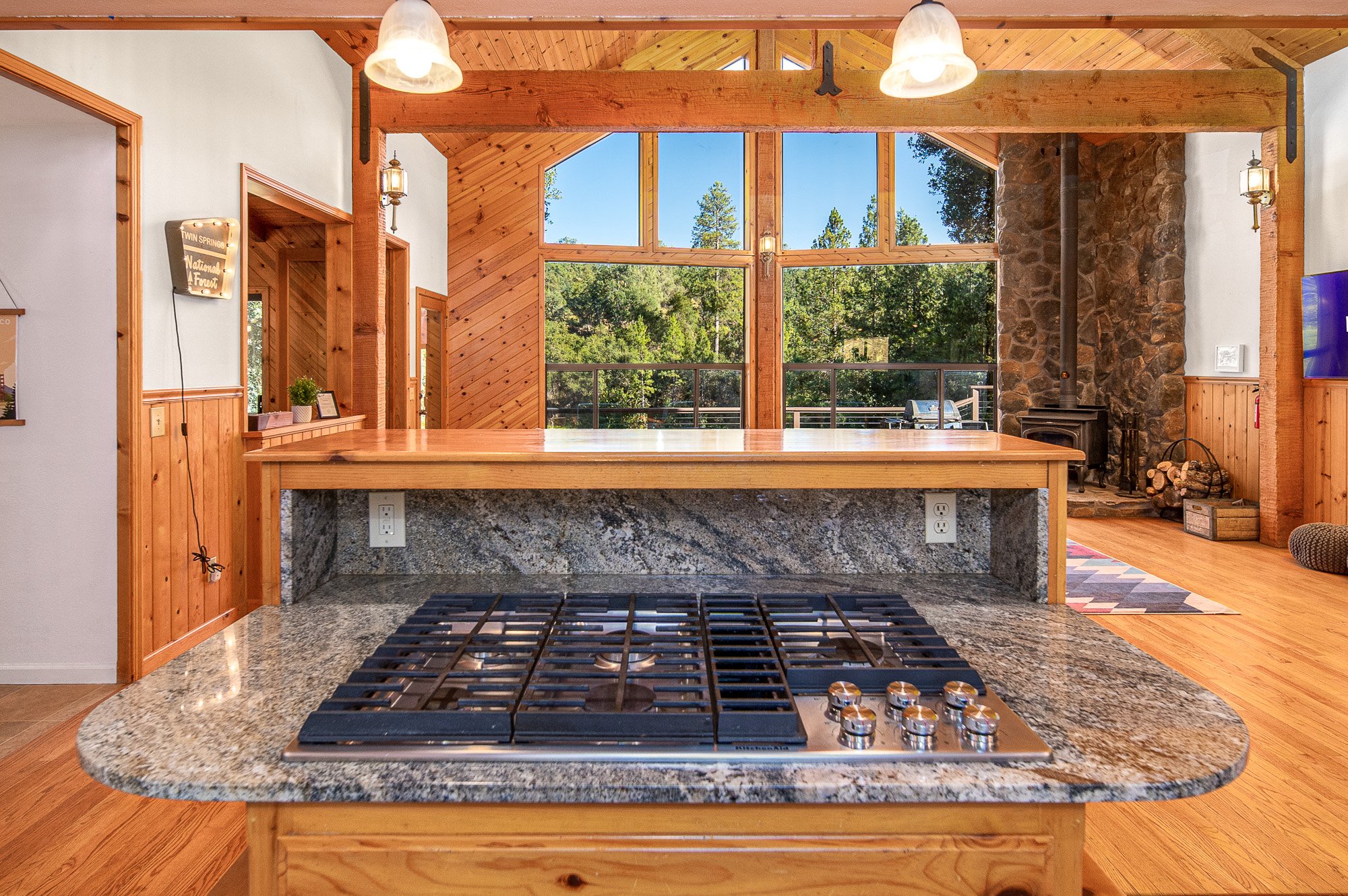 Rustic kitchen interior with gas stove on granite countertop, wooden cabinetry, large windows with forest view, stone fireplace, and wood flooring.