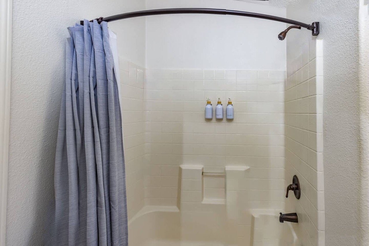 Bathroom shower with a curved curtain rod, blue shower curtain, and three wall-mounted dispensers against white tiled walls.