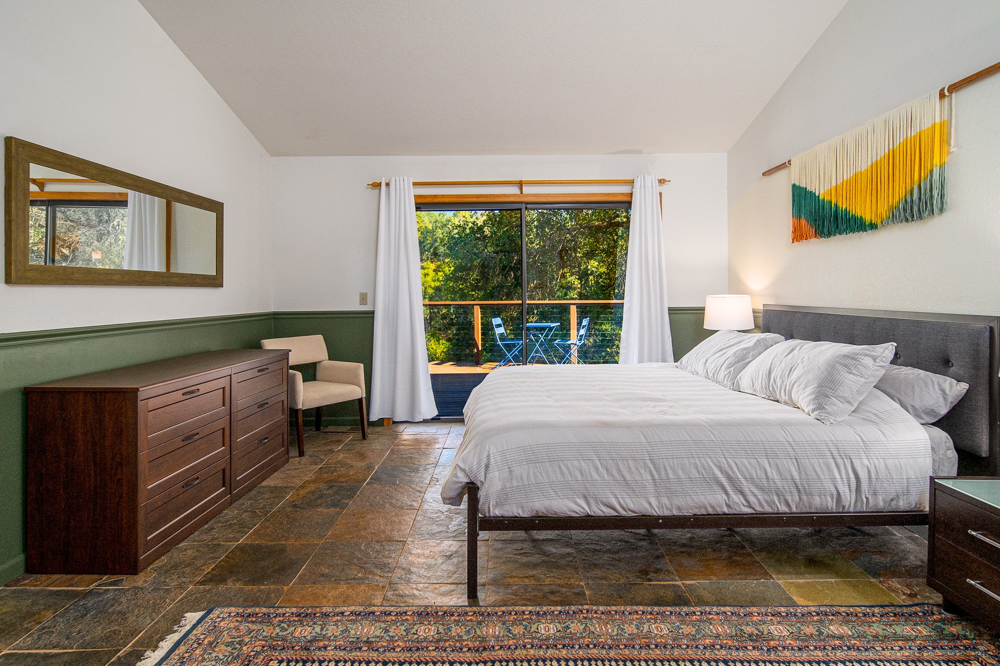 Modern bedroom with a large bed, white linens, wooden dresser, woven wall art, and a sliding glass door opening to a patio with outdoor furniture and a view of trees.