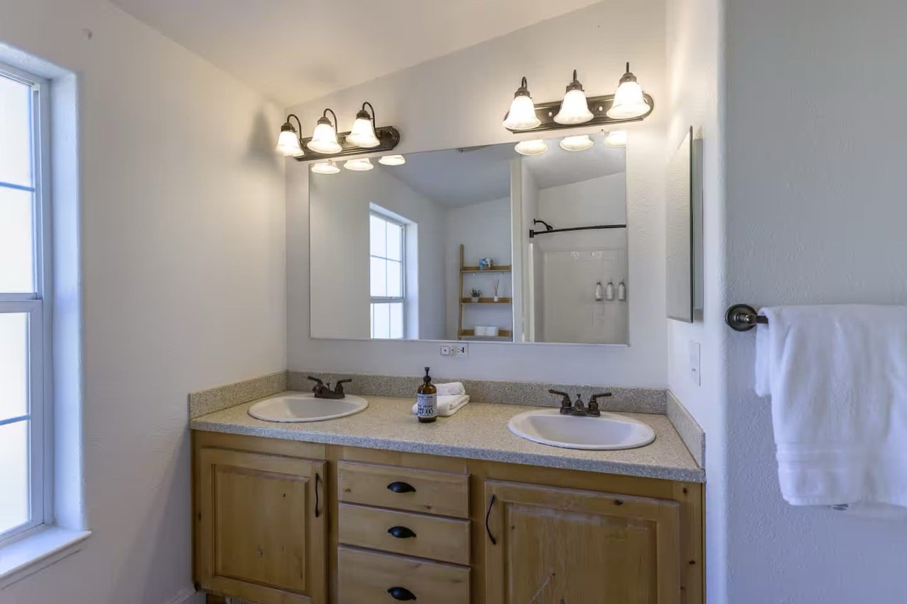 Modern bathroom with double sinks, large mirror, wooden cabinets, countertop toiletries, overhead lighting, shower, and towel rack.