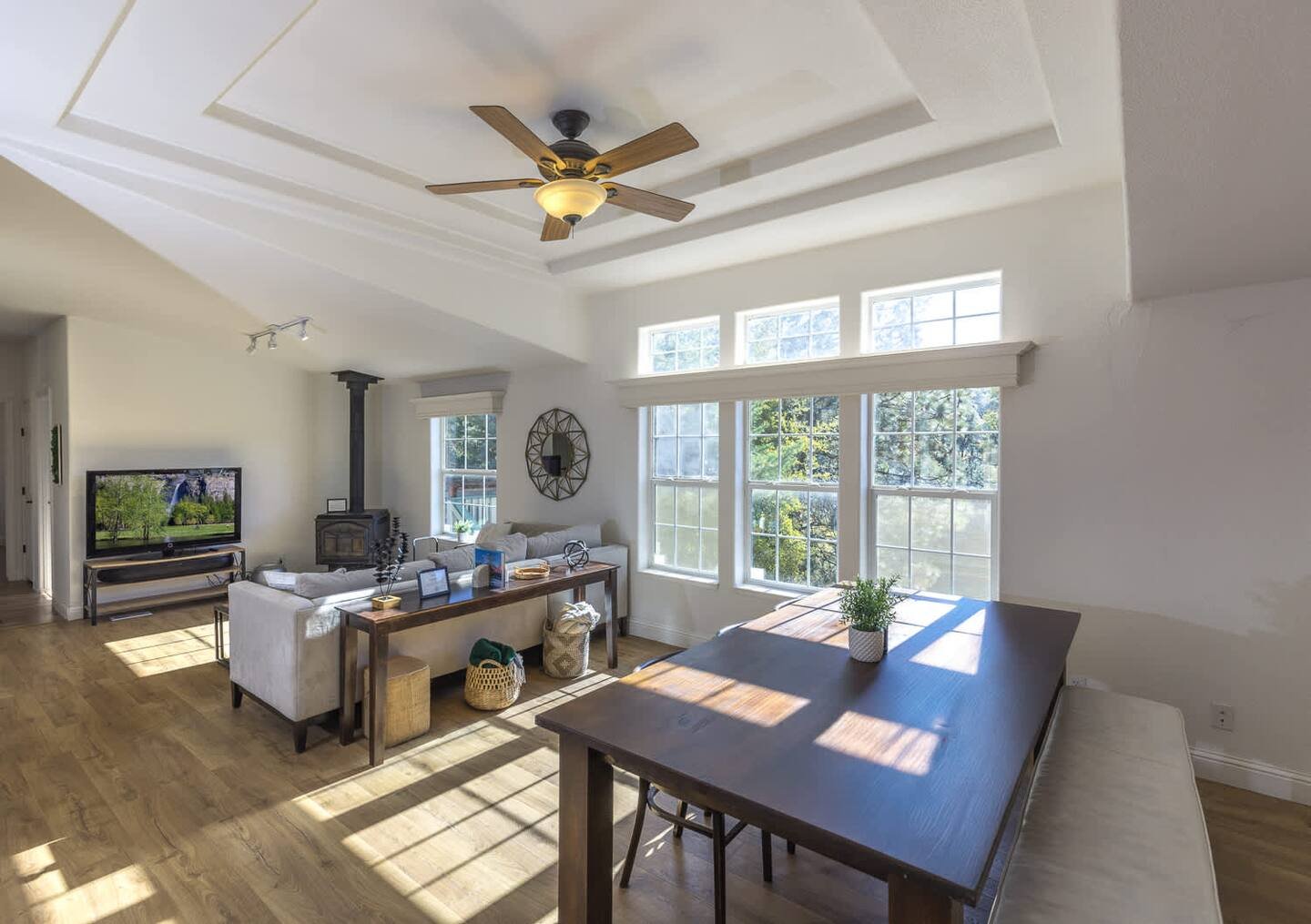 Bright living room interior with a ceiling fan, wooden floor, and large windows. Features a dining table, TV, couch, and decorative items. Natural light illuminates the space.