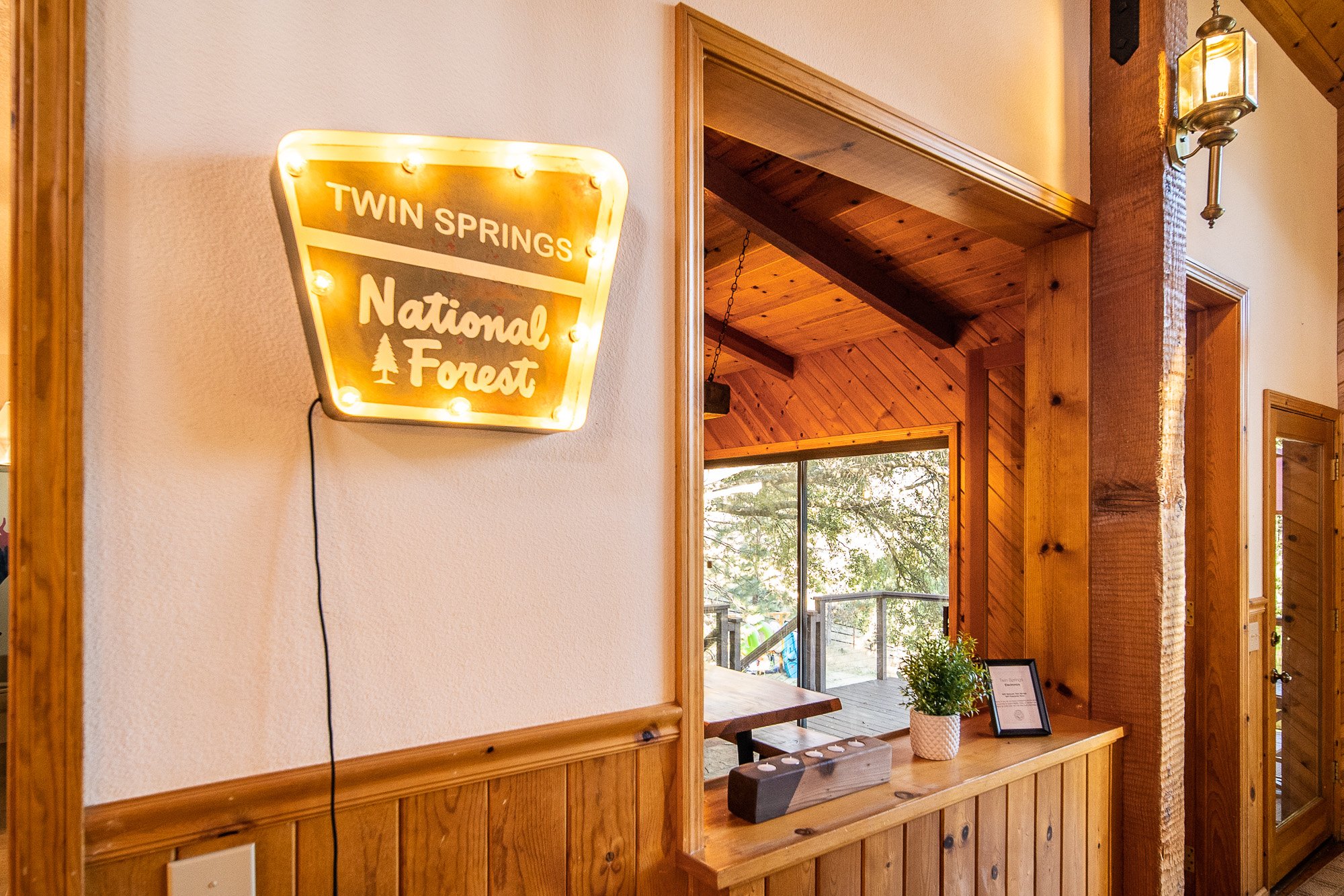 Interior view of a rustic wooden cabin featuring a lit "Twin Springs National Forest" sign, wooden paneling, a potted plant, and a large window displaying an outdoor deck with trees.