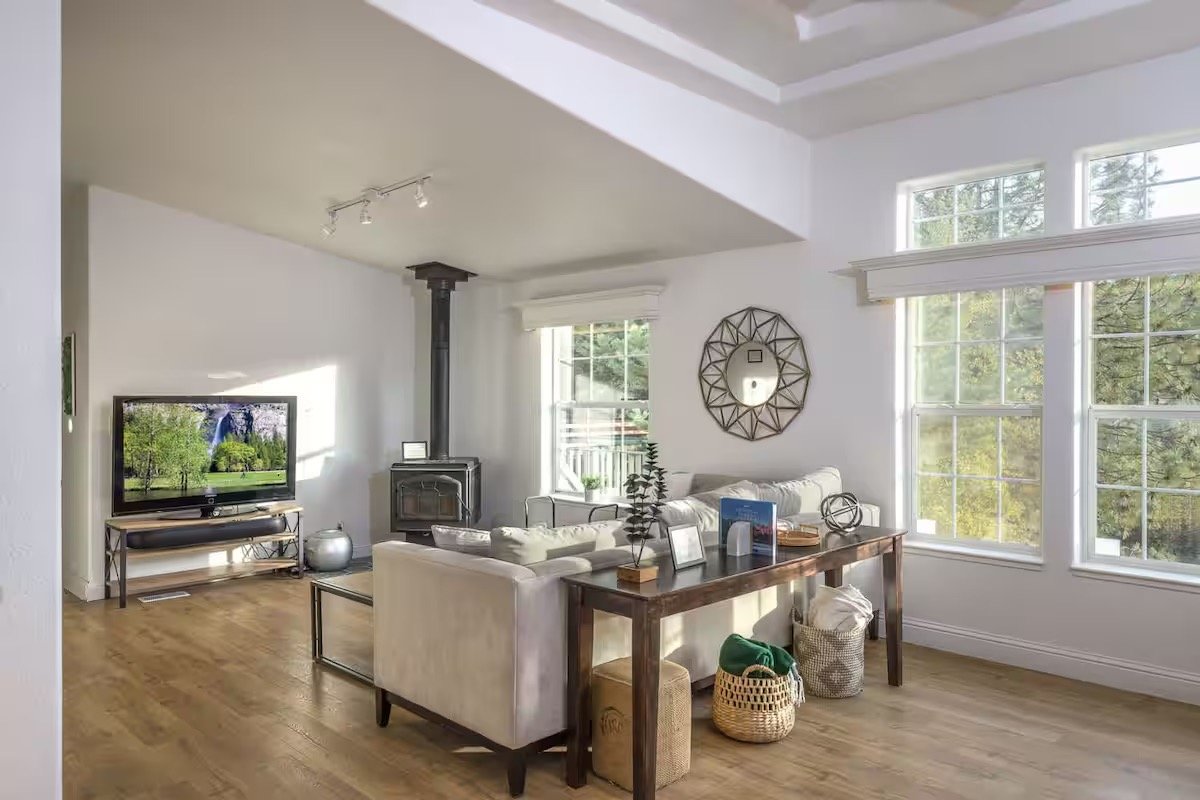 Bright living room with a large window, cozy gray couch, wood-burning stove, wall-mounted TV, and decorative items on a wooden table.