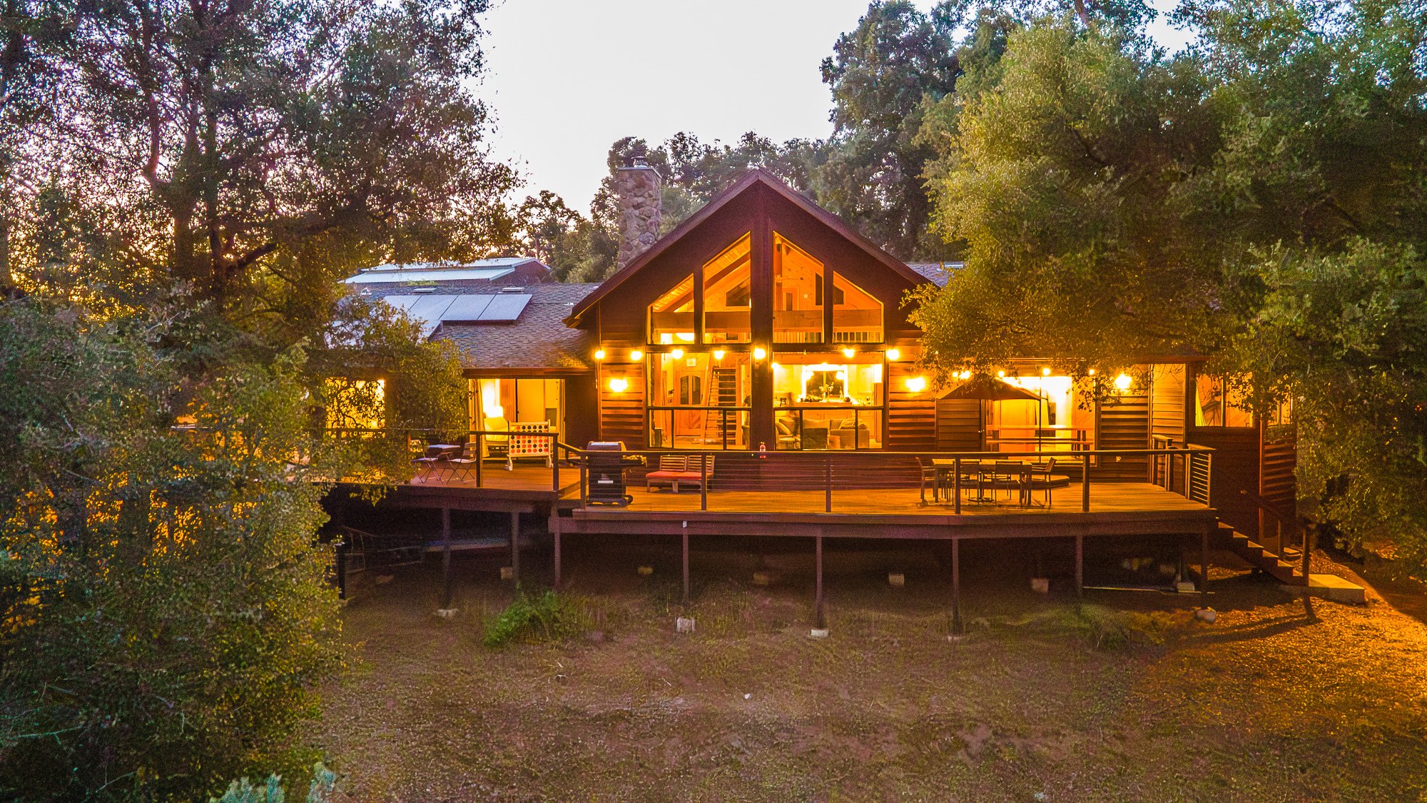 A wooden cabin with large glass windows and a spacious deck, surrounded by trees. The cabin is illuminated with warm lights and features outdoor furniture on the deck.