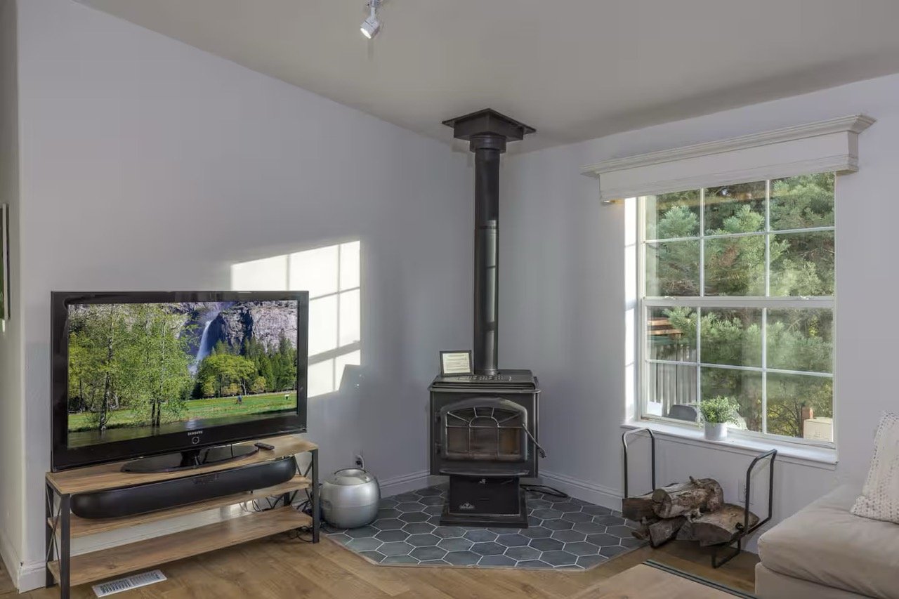 Living room with a TV on a stand, wood-burning stove, logs in a holder, and large window with outdoor view.