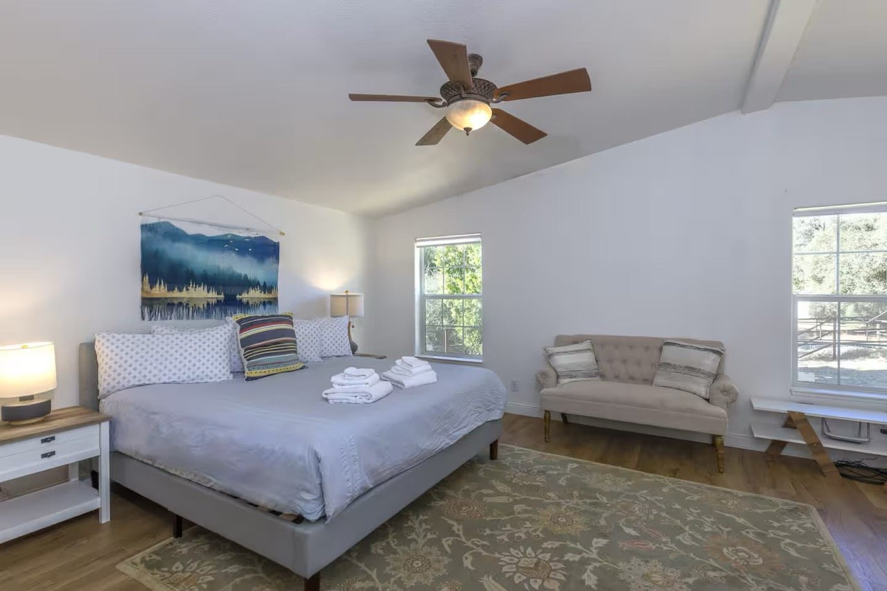 Spacious bedroom with a king-sized bed, decorative pillows, folded towels, nightstands with lamps, wall art, a ceiling fan, a sofa, and large windows with natural light.