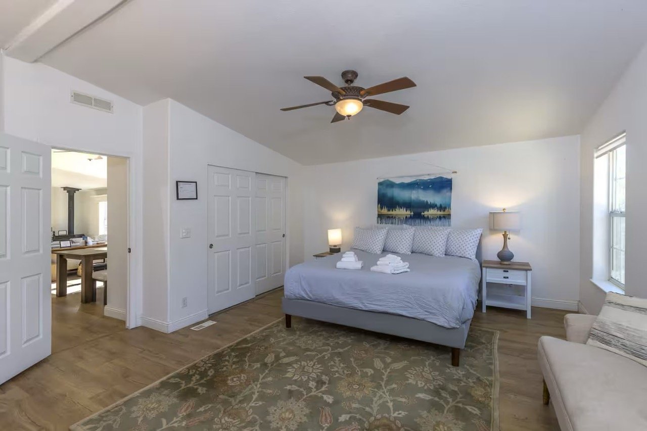 Bedroom with a large bed, white bedding, pillows, an abstract painting above, a ceiling fan, wood flooring, lamp on a nightstand, a decorative rug, and a partially open door leading to another room.