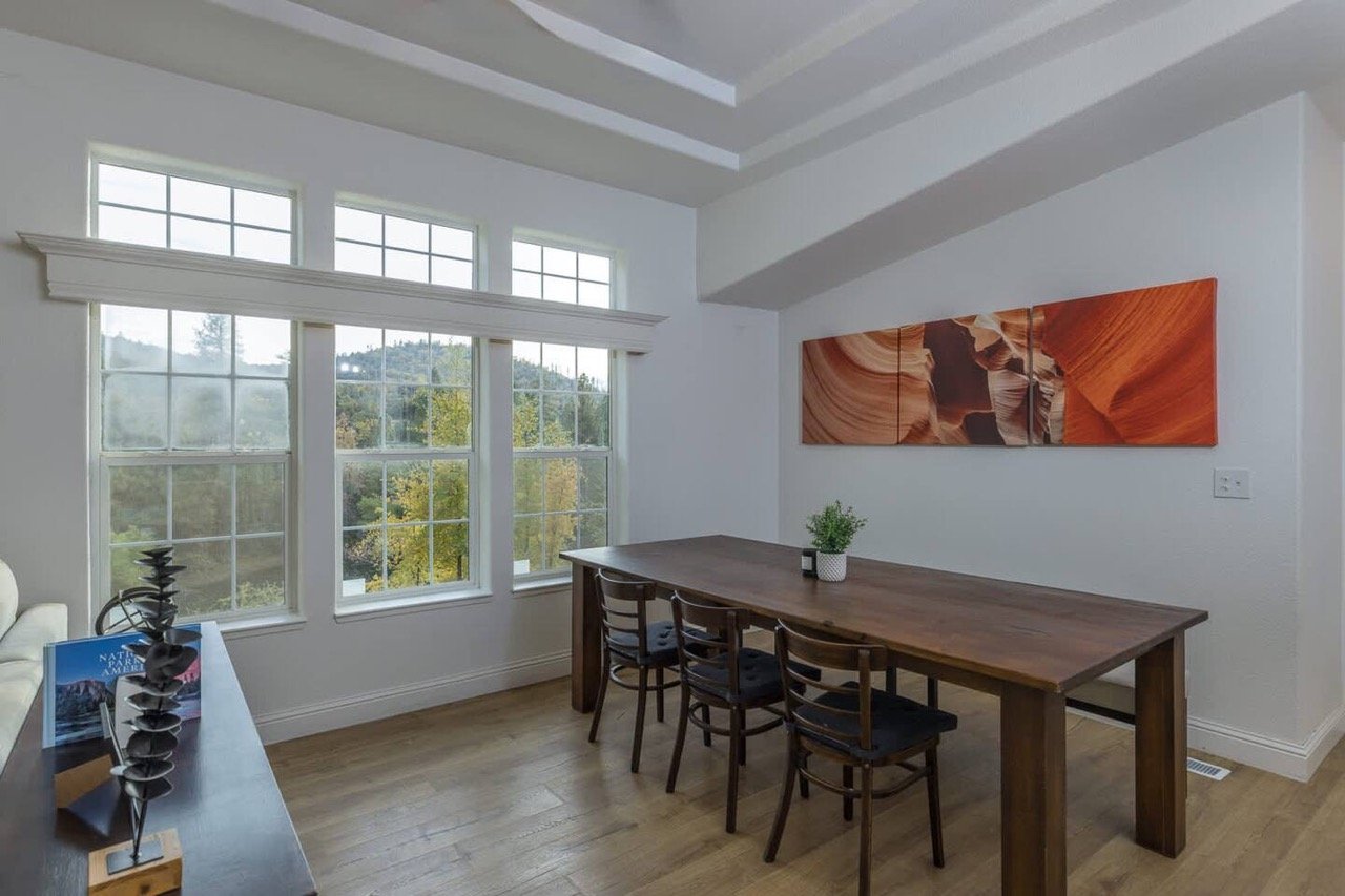 Minimalist dining room with wooden table, four chairs, landscape artwork, and large windows overlooking a scenic view.