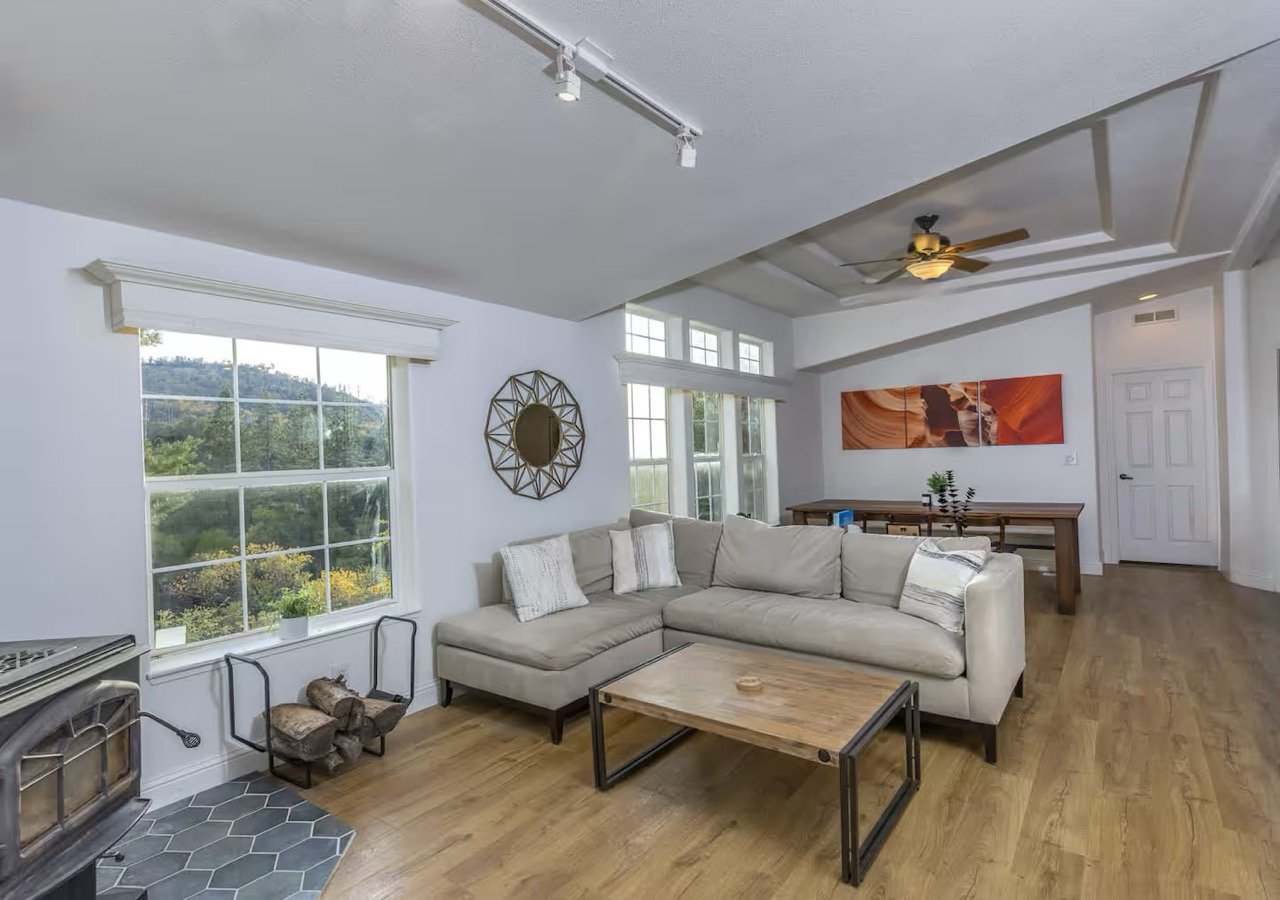 Modern living room with sectional sofa, wooden coffee table, large windows, wall art, ceiling fan, and a wood-burning stove with a log holder.