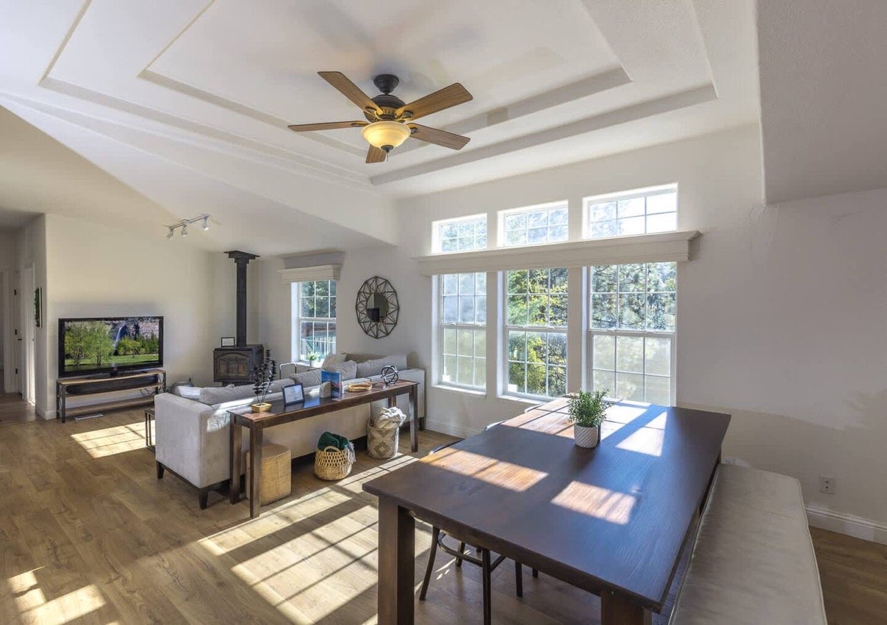 Bright living and dining area with wood floors, featuring a ceiling fan, large windows, a dining table, sofas, a wall-mounted TV, and a wood-burning stove.