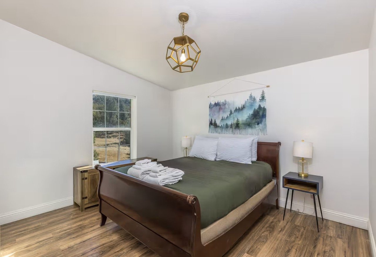 Minimalist bedroom with wooden bed, forest-themed wall art, and modern lighting.