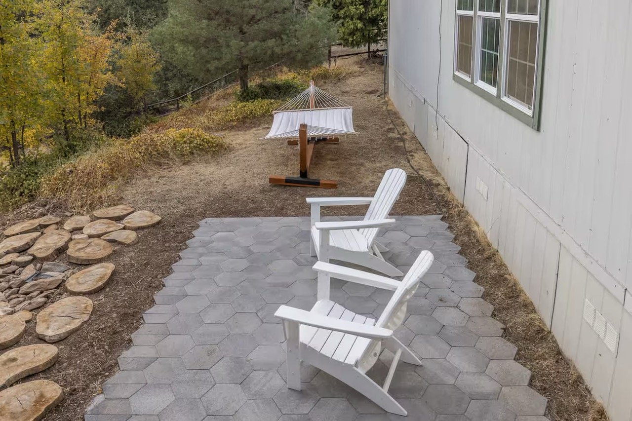 Outdoor patio with two white Adirondack chairs and a hammock on a stand, surrounded by trees and a stone pathway.