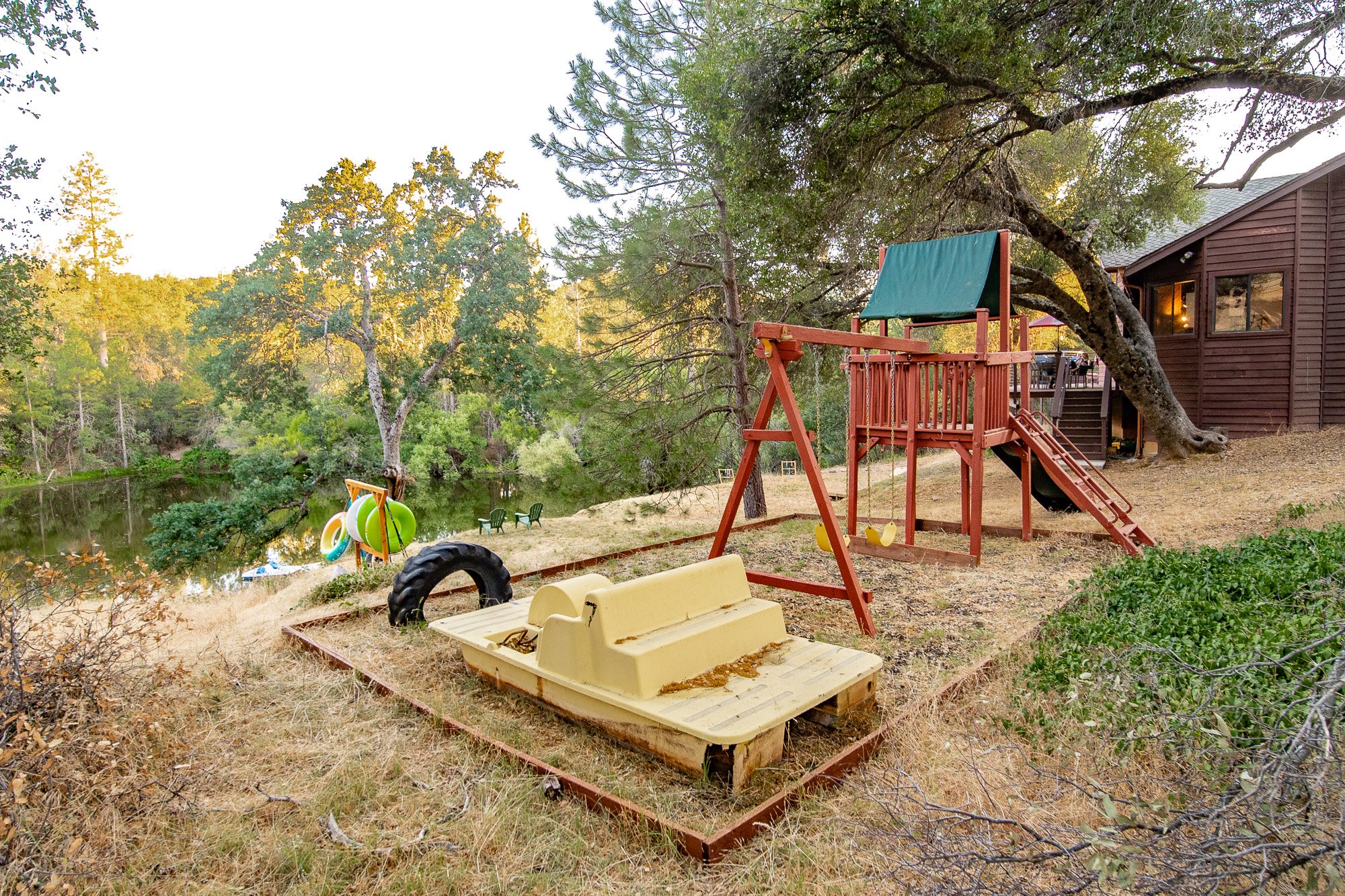 Backyard with wooden playset, slide, swing, and rubber tire in dry grassy area next to a brown house with trees and lake view.