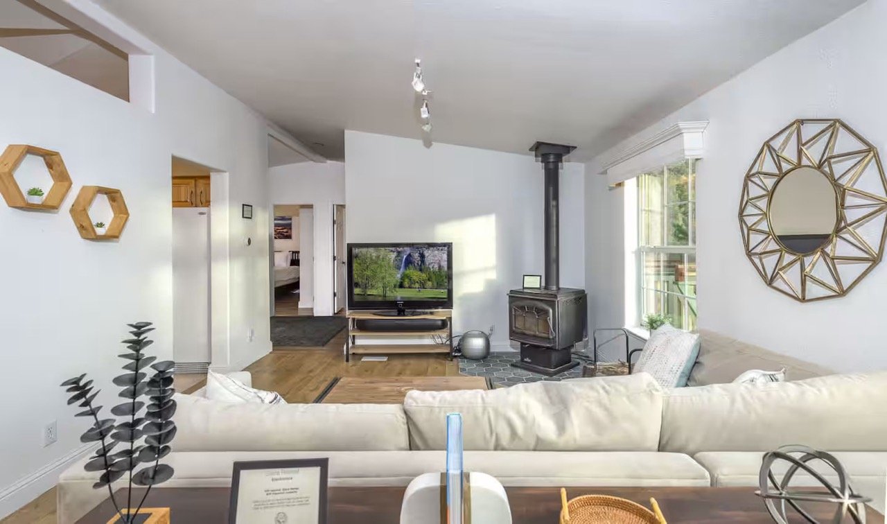 Living room with a white couch, TV on a stand, wood-burning stove, geometric mirror, and hexagonal wall shelves.