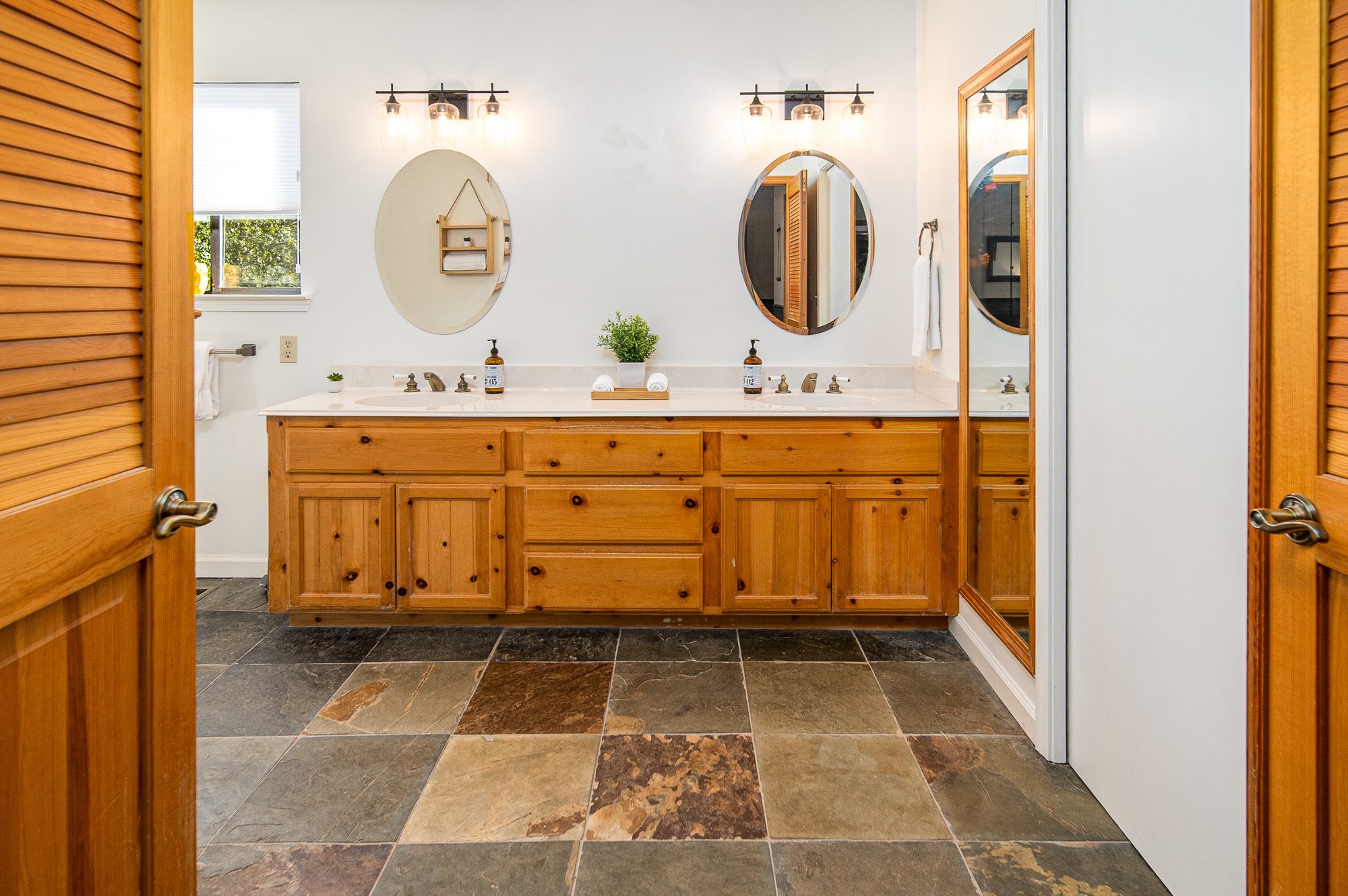 Bathroom with wooden cabinets, dual sinks, oval mirrors, hexagon tiled floor, and glass light fixtures.