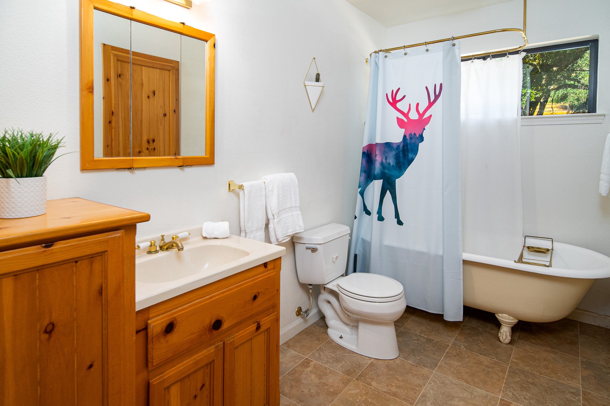 Bathroom with wooden vanity, mirror, toilet, clawfoot bathtub, and shower curtain with colorful deer design.