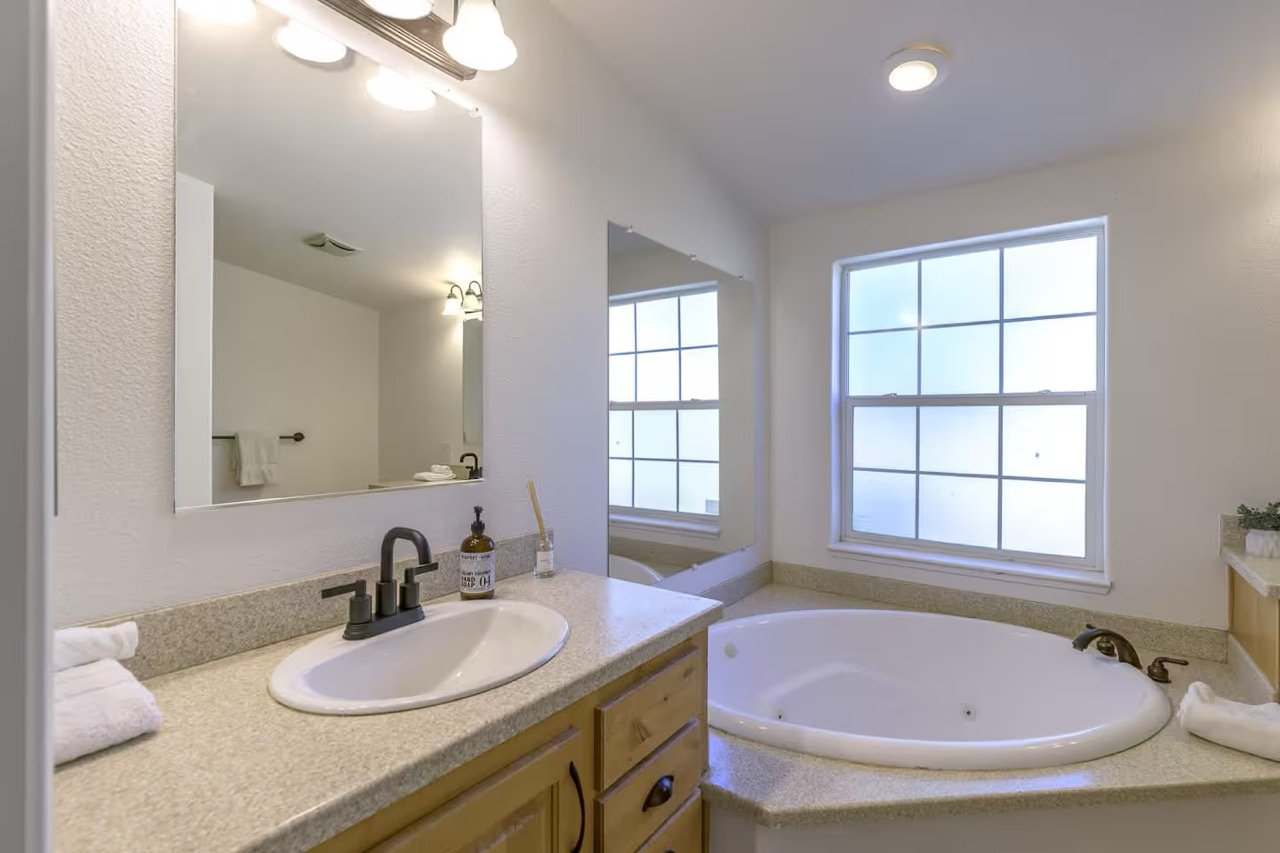 Bathroom with a vanity sink, large mirror, and corner bathtub under a window.