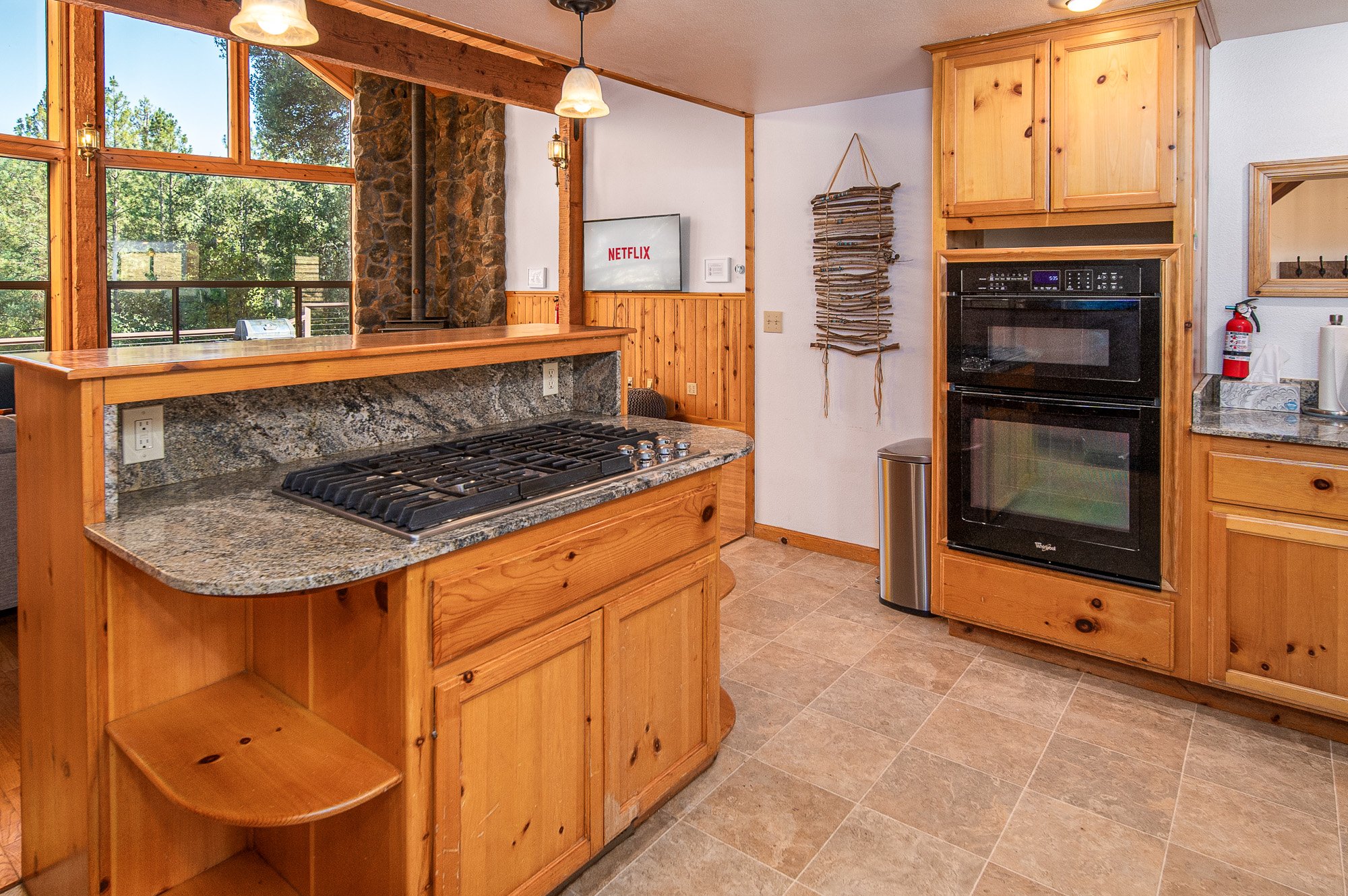 Cozy kitchen with granite countertops, wooden cabinets, gas stove, double oven, and TV on wall. Large windows offer forest view. Decorative wall hanging and fire extinguisher visible.