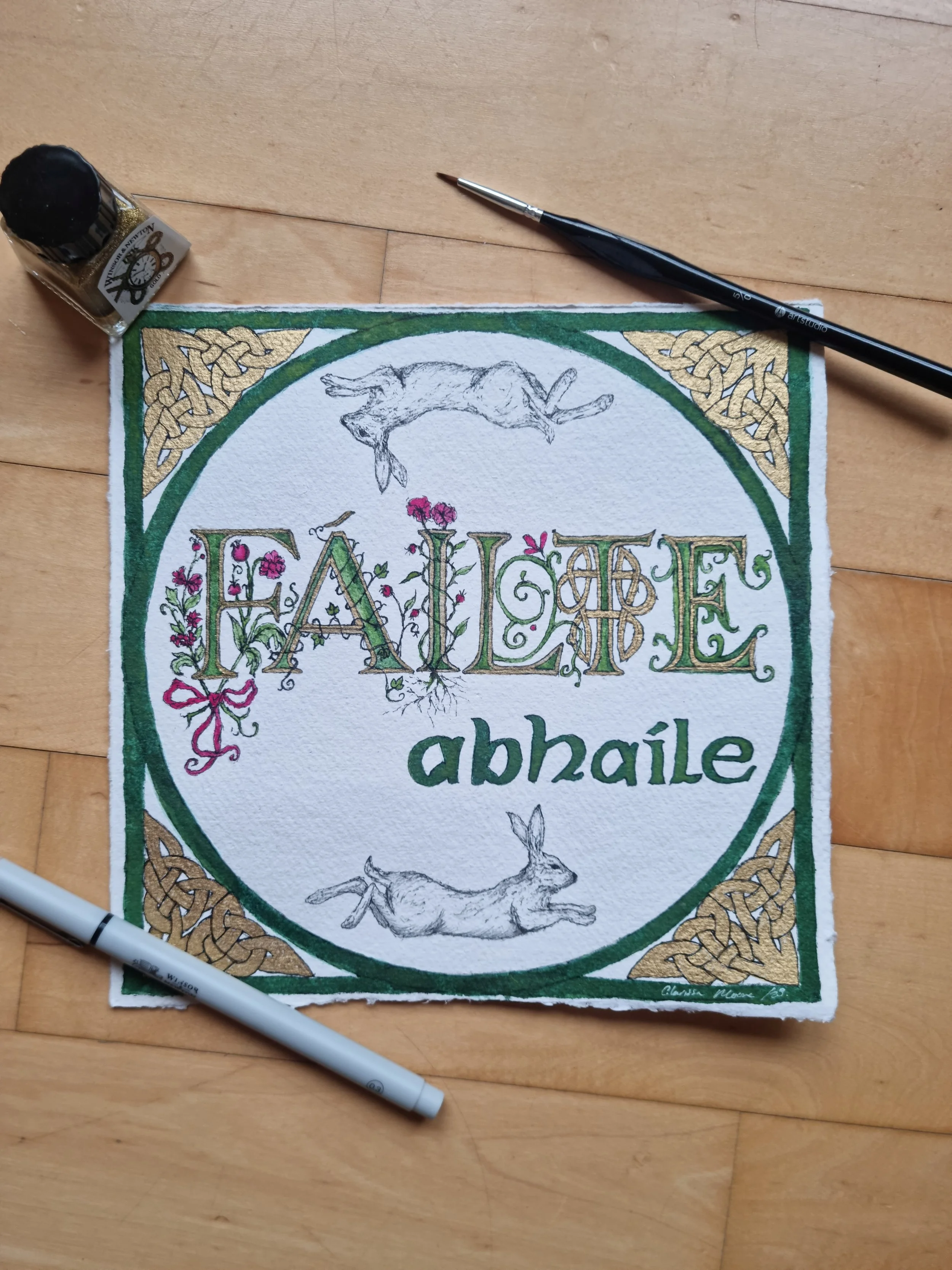 A partially colored Irish-themed artwork with the word 'FAILE' in decorative letters, the word 'abhaile' below, illustrations of rabbits and a dog, and Celtic knots in the corners, surrounded by art supplies on a wooden surface.