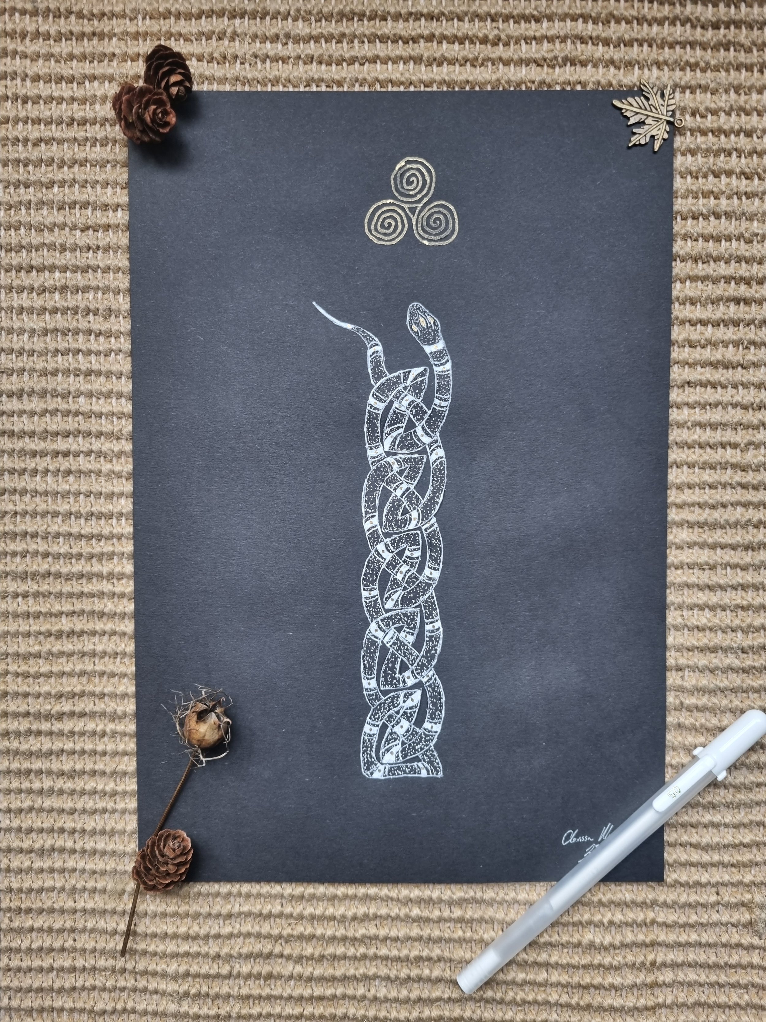 White illustration of a serpent intertwined with Celtic knots on black paper, decorated with pinecones, a dried seed pod, a leaf-shaped pin, and a white marker.