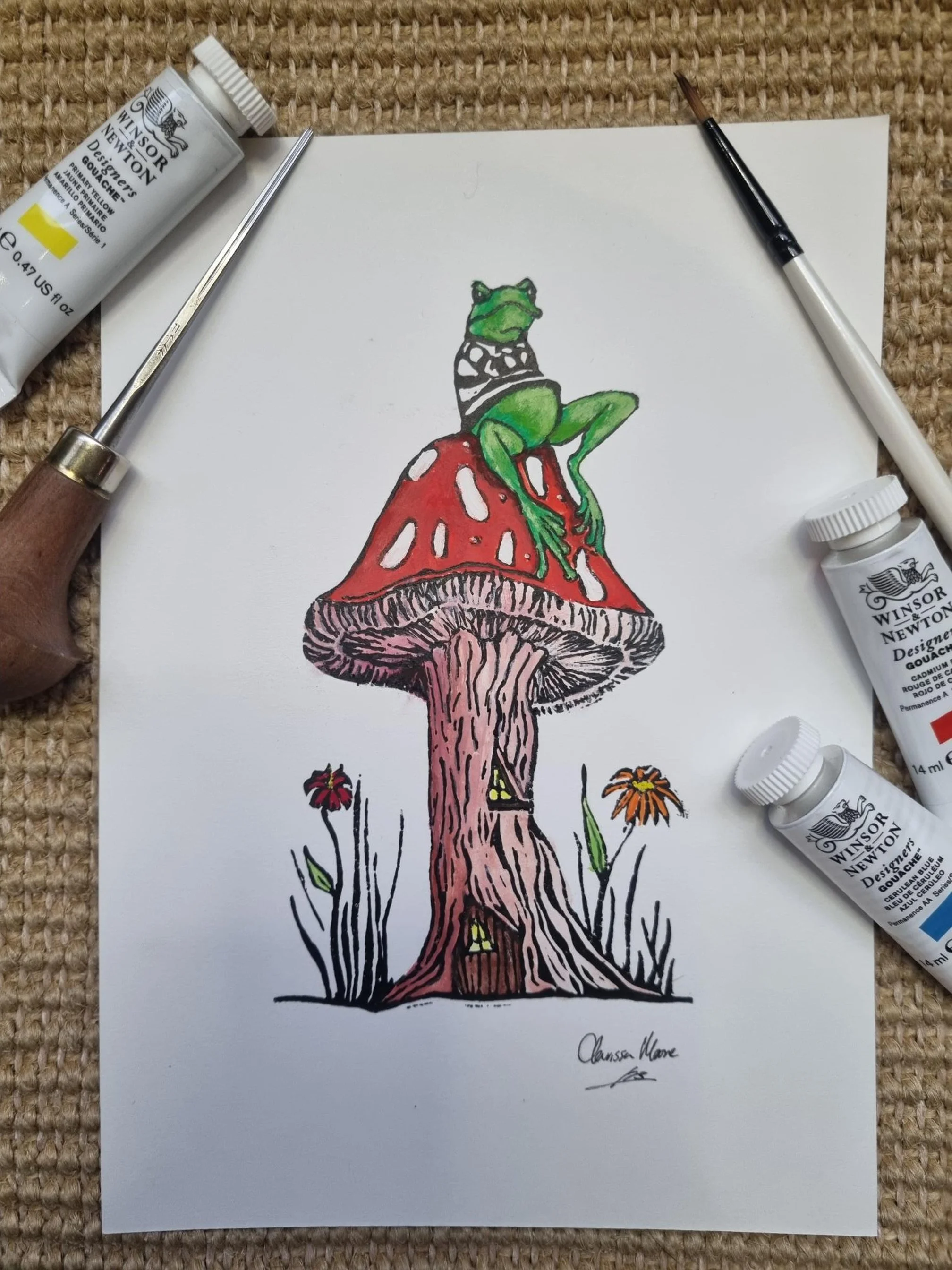 A colorful drawing of a frog sitting on a red and white mushroom, with a smaller mushroom and flowers at the base. Art supplies, including paint tubes, brushes, and a palette knife, surround the drawing.