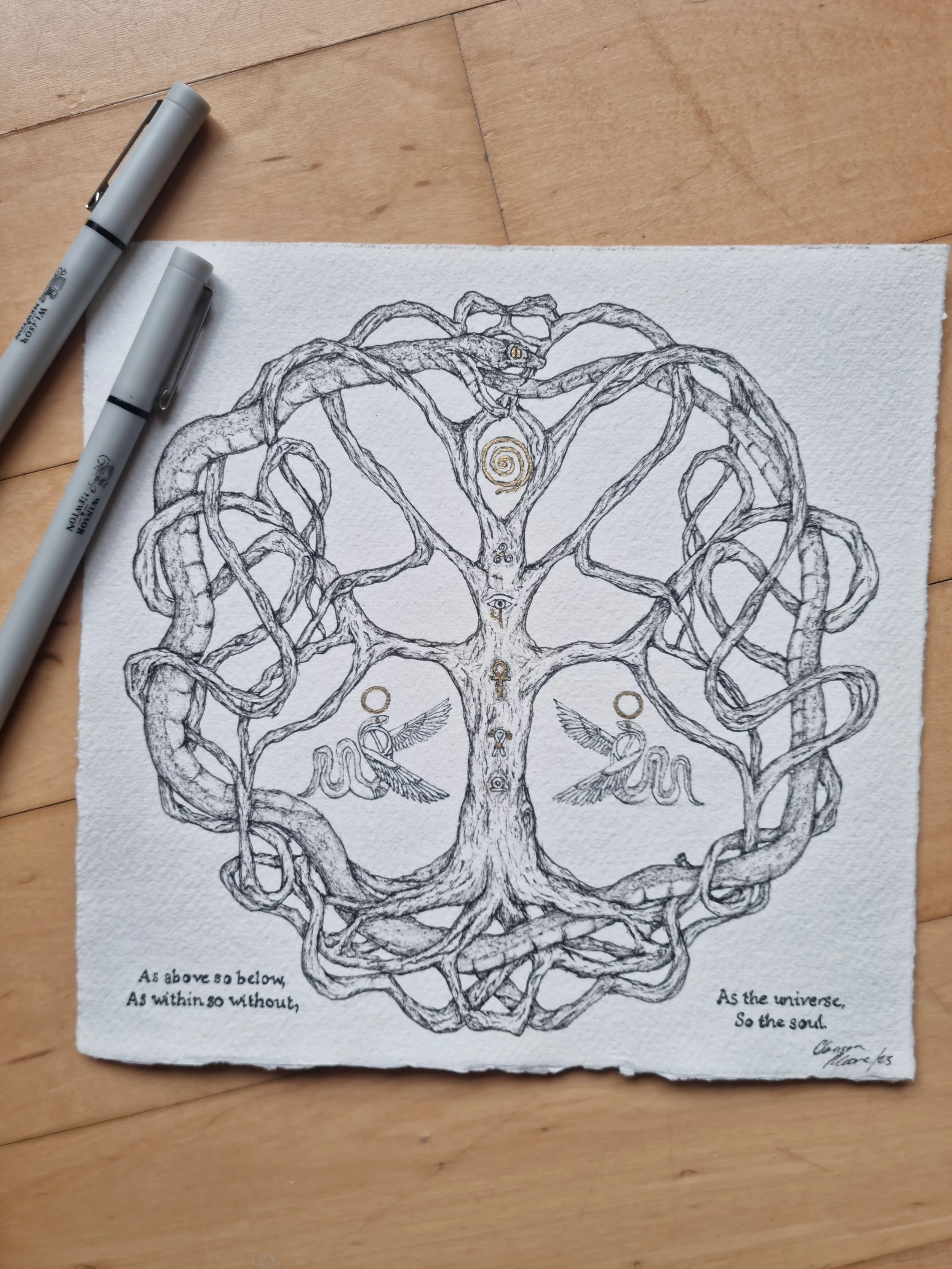 A detailed drawing of a tree with intertwined branches, forming a circular shape. The tree has various mystical symbols, including an eye, a spiral, an ankh, and a winged snake. There is a quote at the bottom left and right corners. Two gray pens are placed beside the drawing on a wooden surface.