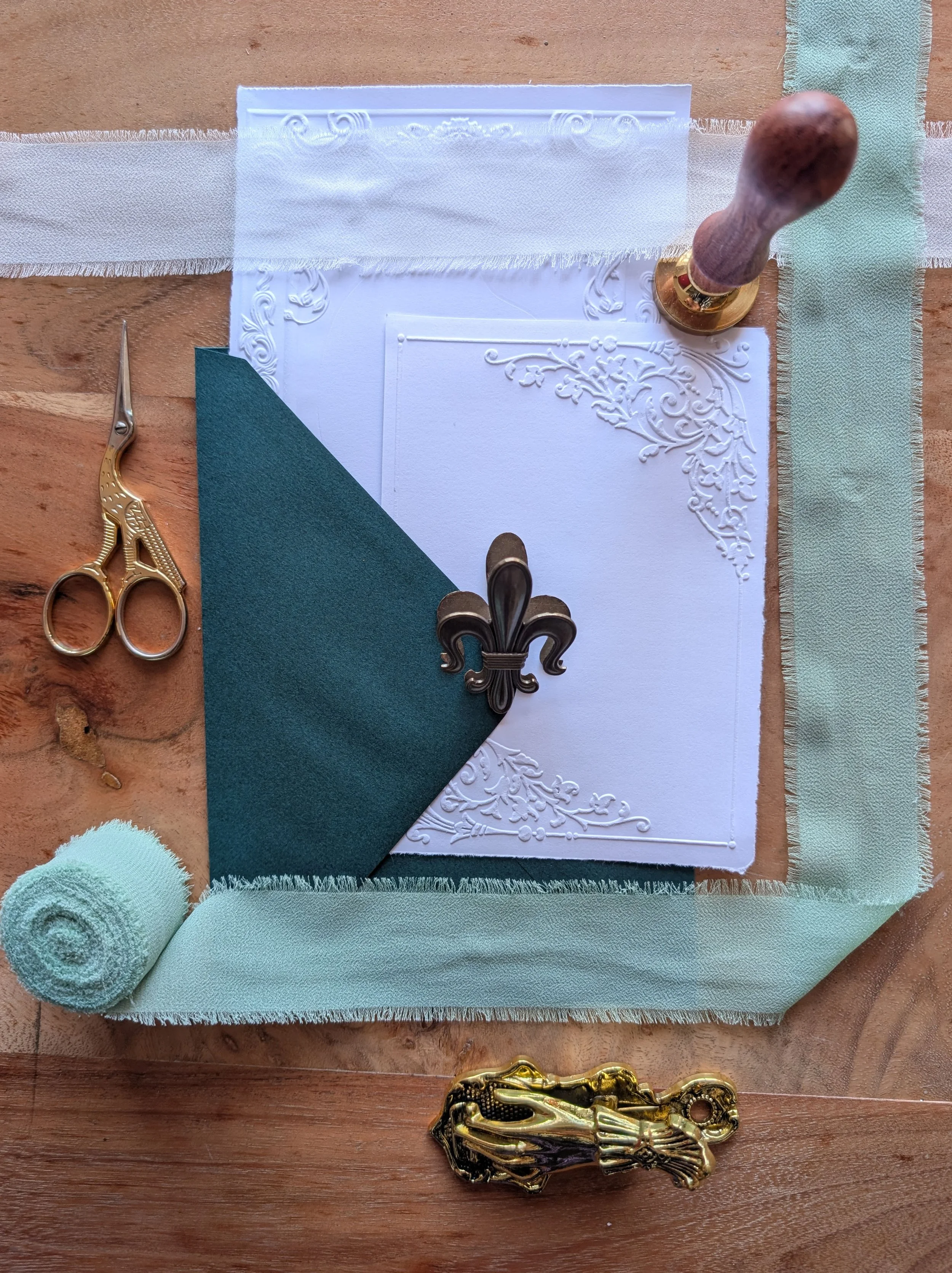 Flat lay of decorative items and craft supplies, including scissors, a fleur-de-lis pin, a rubber stamp, a rolled towel, and fabric strips on a wooden surface, with lace and embossed paper as background.