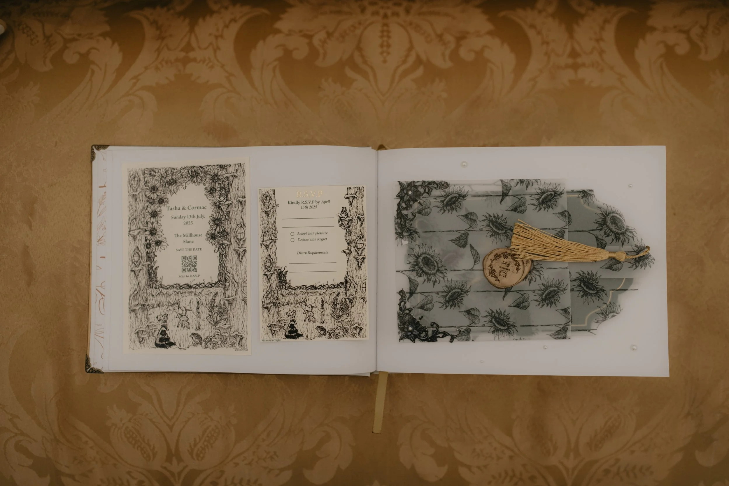 Open book displaying black and white illustrations and stationery, including wedding invitations with floral designs and a wooden tag with a gold tassel on top of a wrapped gift box with sunflower patterns, on a gold-patterned surface.