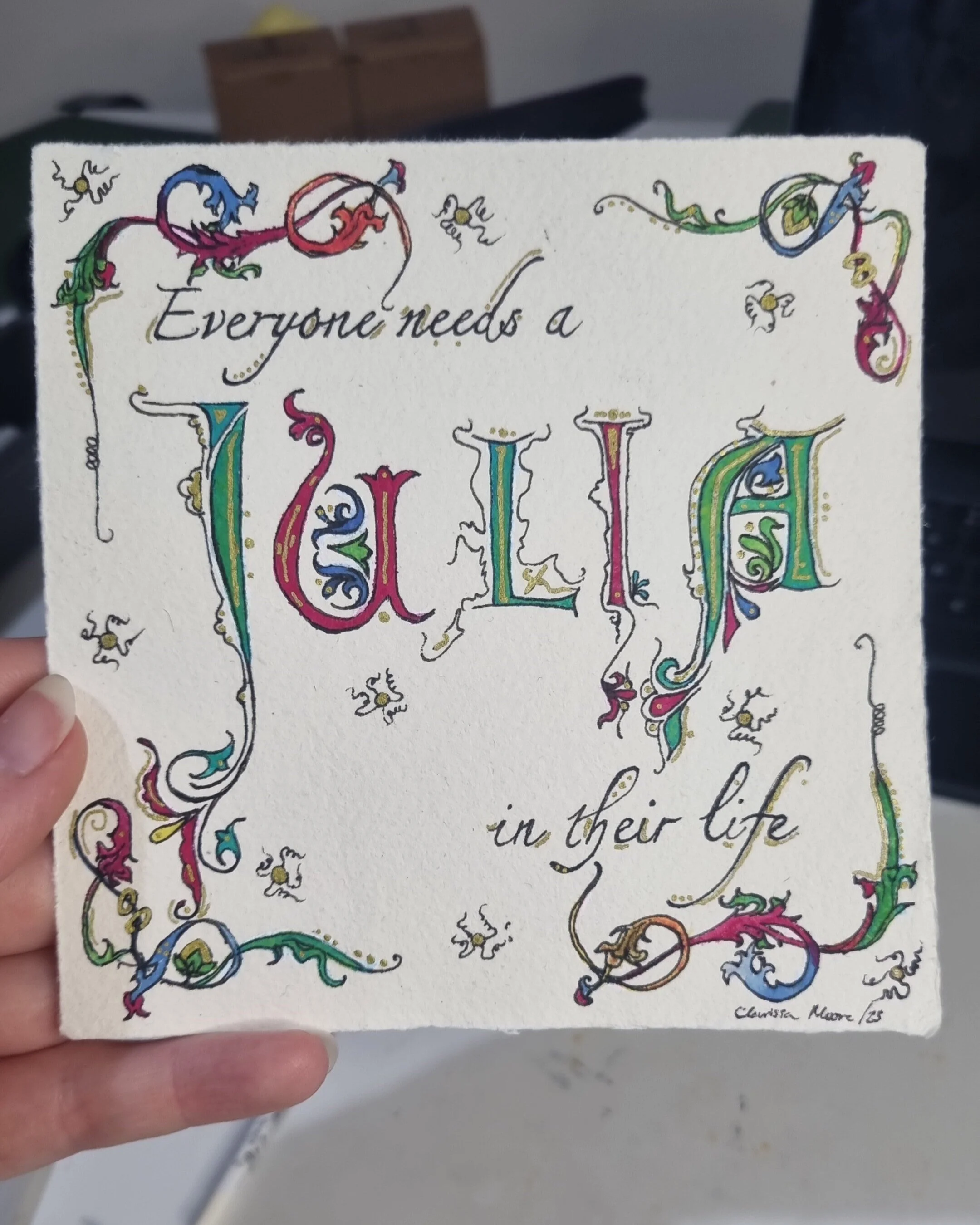 Decorative illustrated quote on a white card that says, "Everyone needs a little in their life," with colorful, ornate lettering and small bees and floral designs surrounding the text.
