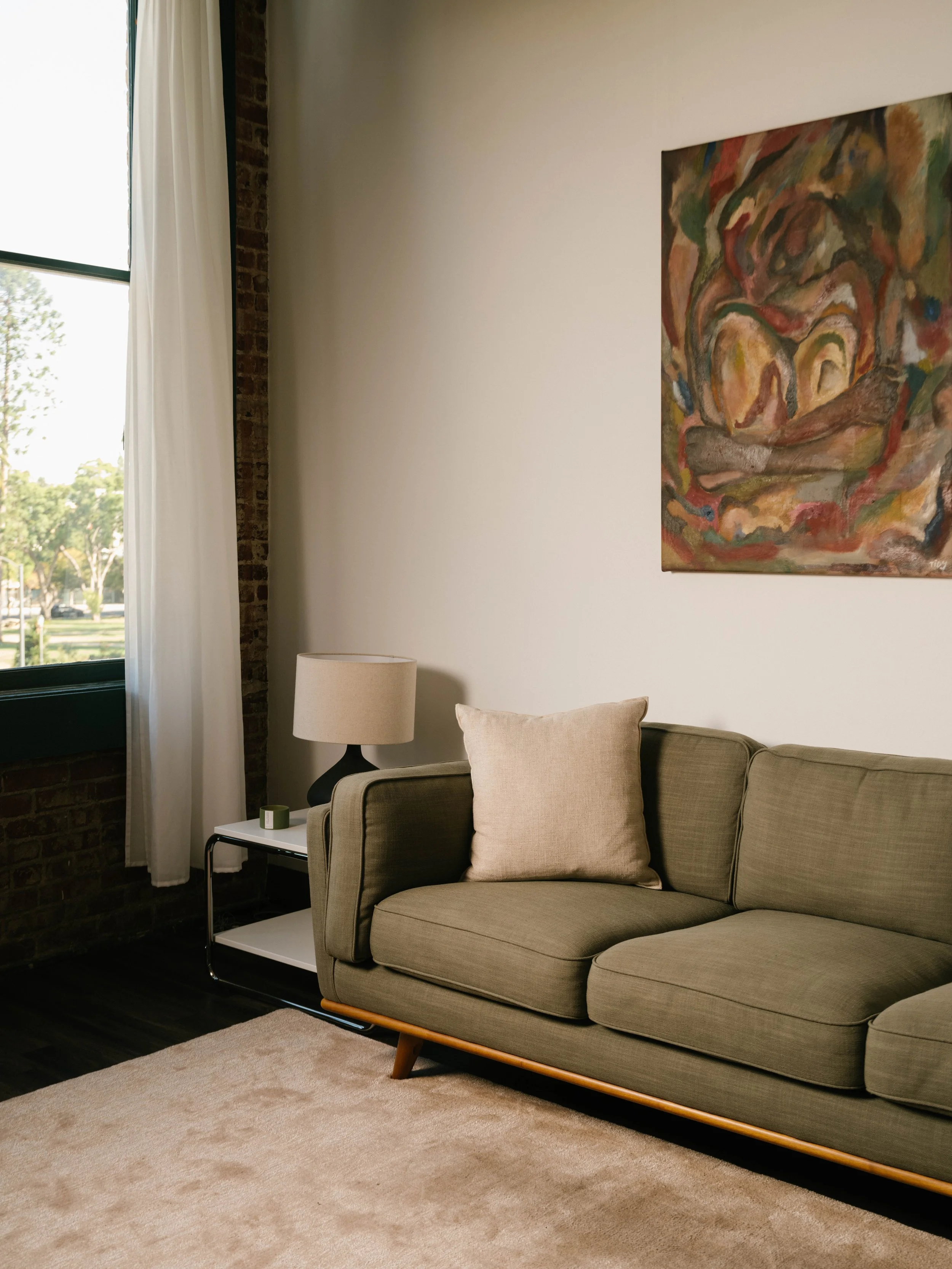Living room corner with a beige sofa, a side table with a lamp, a painting on the wall, a window with white curtains, and a beige carpet.