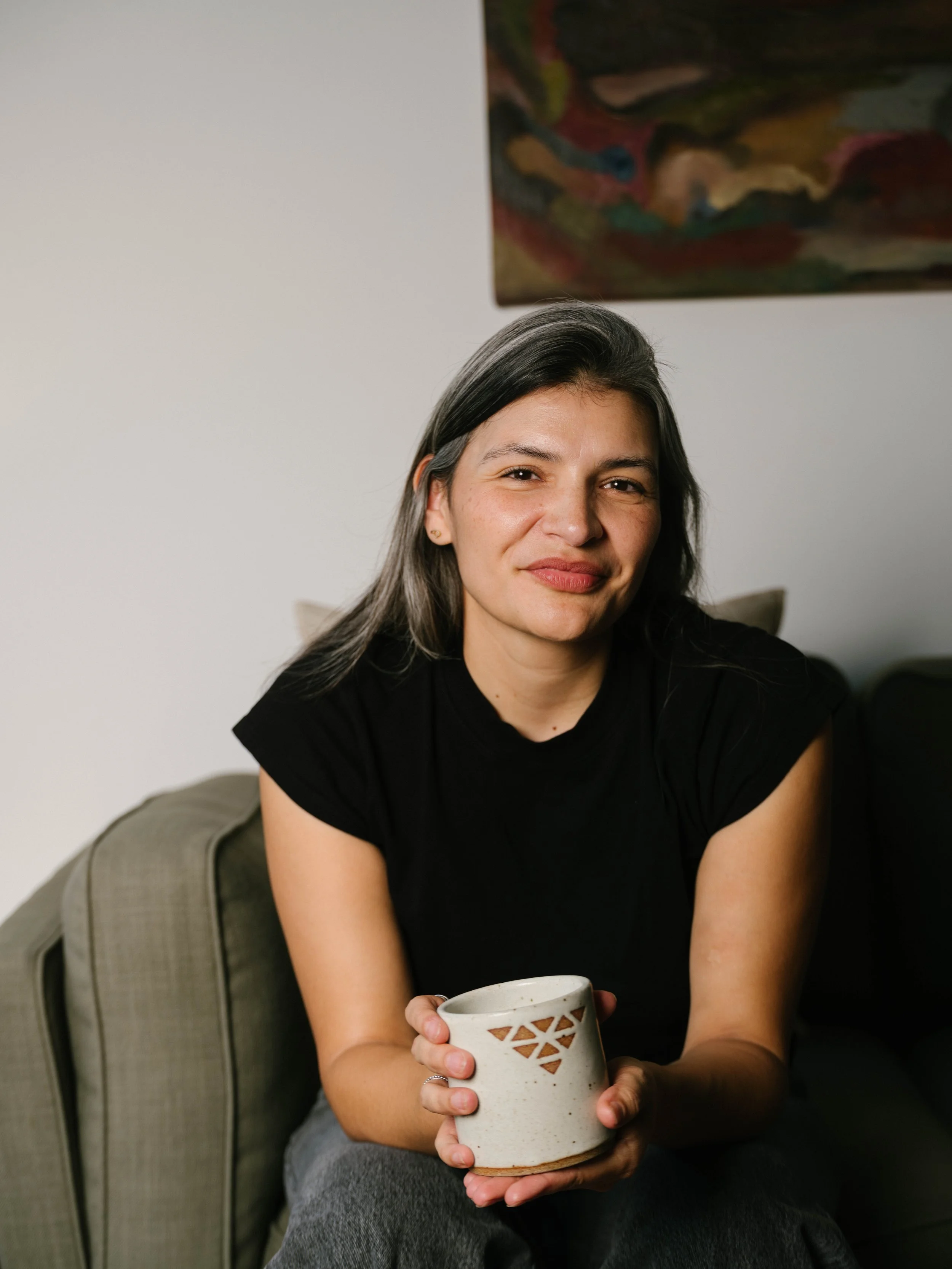 Woman with dark hair and a black shirt sitting on a light green couch, holding a ceramic mug with triangle cutouts, in a room with a colorful abstract painting on the wall.