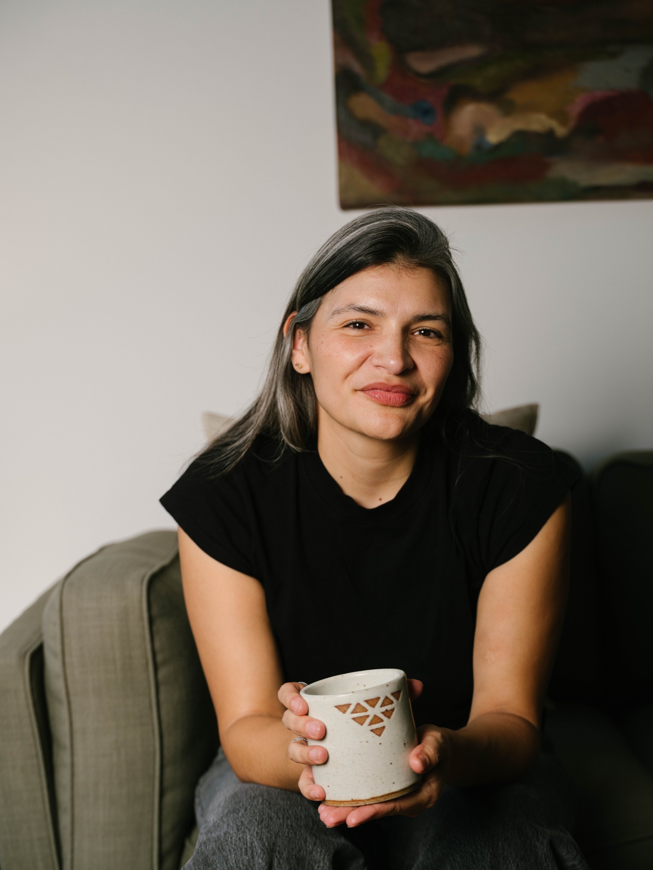 A woman with gray hair, wearing a black shirt, sitting on a beige sofa, holding a white ceramic mug with a geometric cutout design, smiling at the camera.