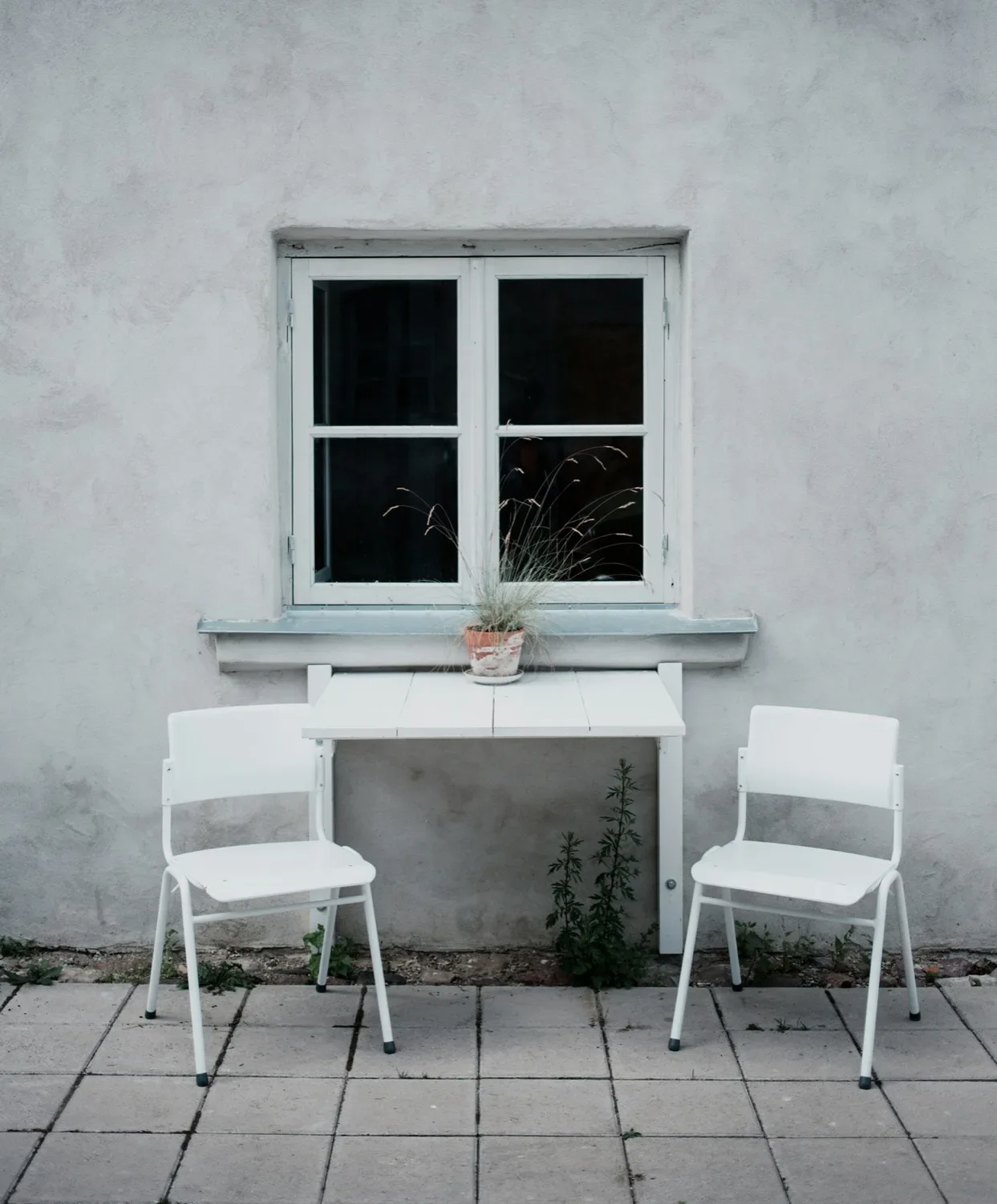 A white outdoor table with a potted plant on top, flanked by two white chairs, placed against a gray stucco wall with a window. The window has a plant behind the glass, and the scene is set on a tiled patio with some small plants growing between the tiles.