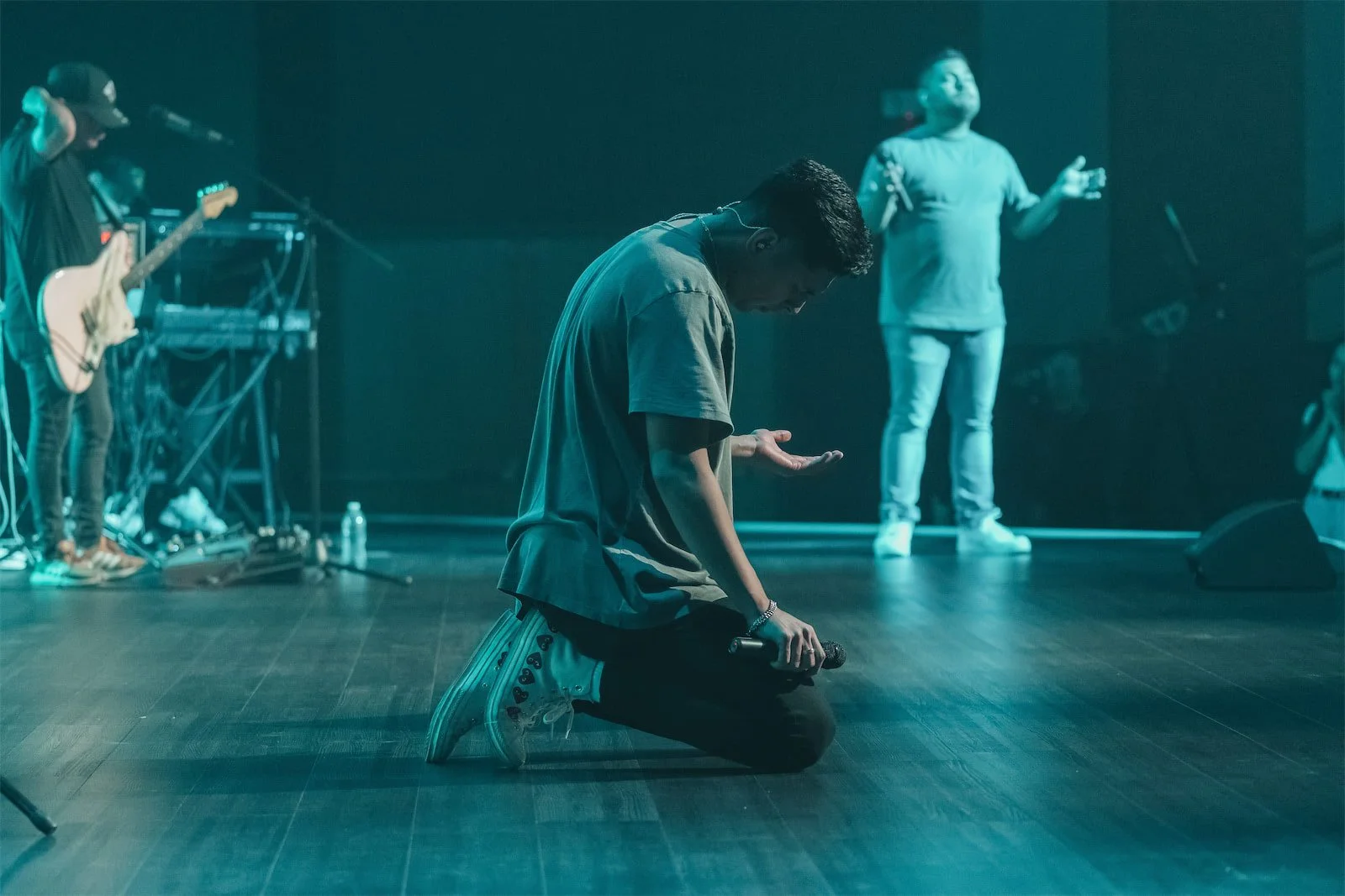 A person kneeling on a stage holding a microphone, with their head bowed and hands raised, while other band members perform in the background.