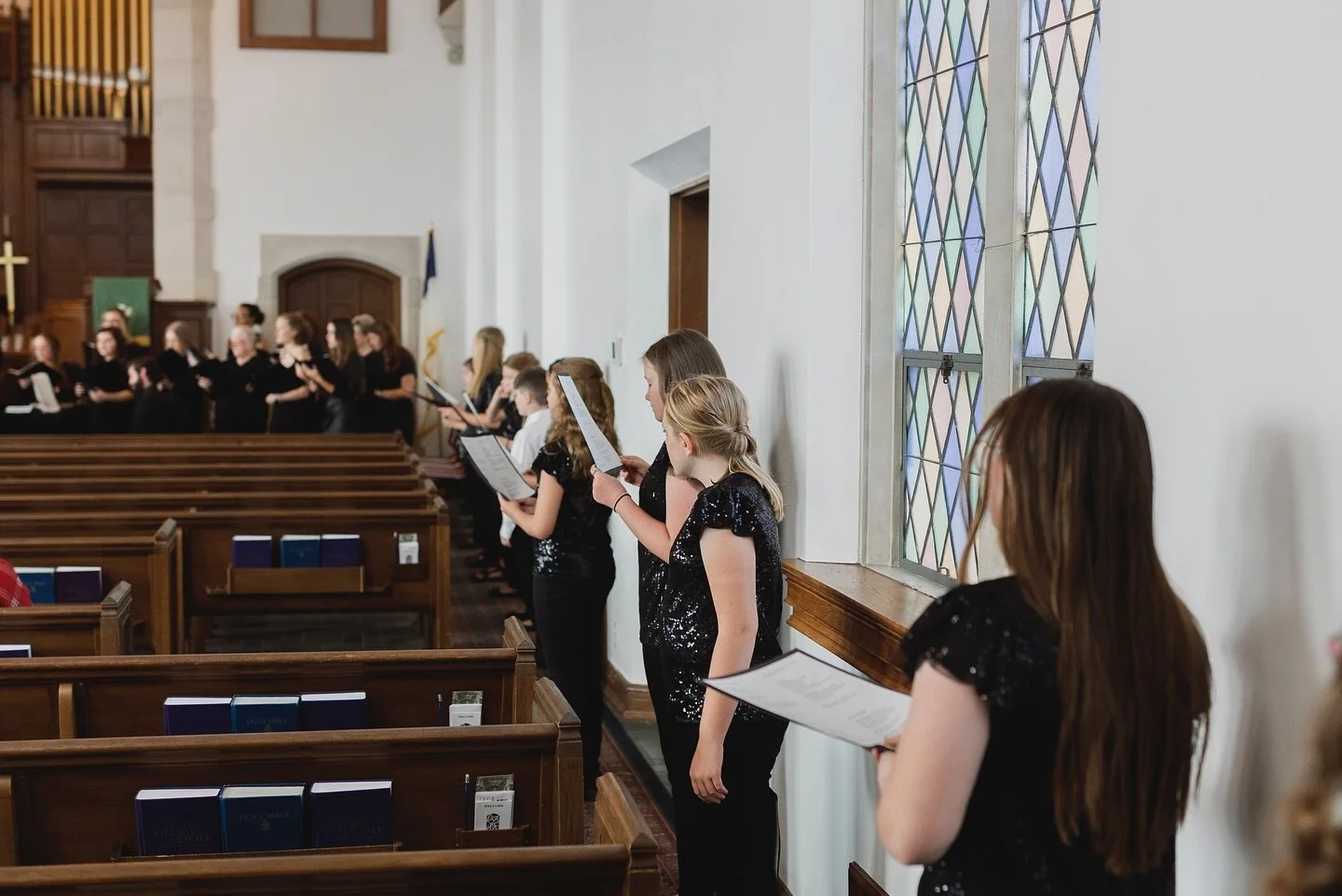 Children and teens singing in a church choir, holding sheet music, wearing black. The choir is facing an altar with a gold cross, stained glass windows are visible on the right, with pews in the foreground.