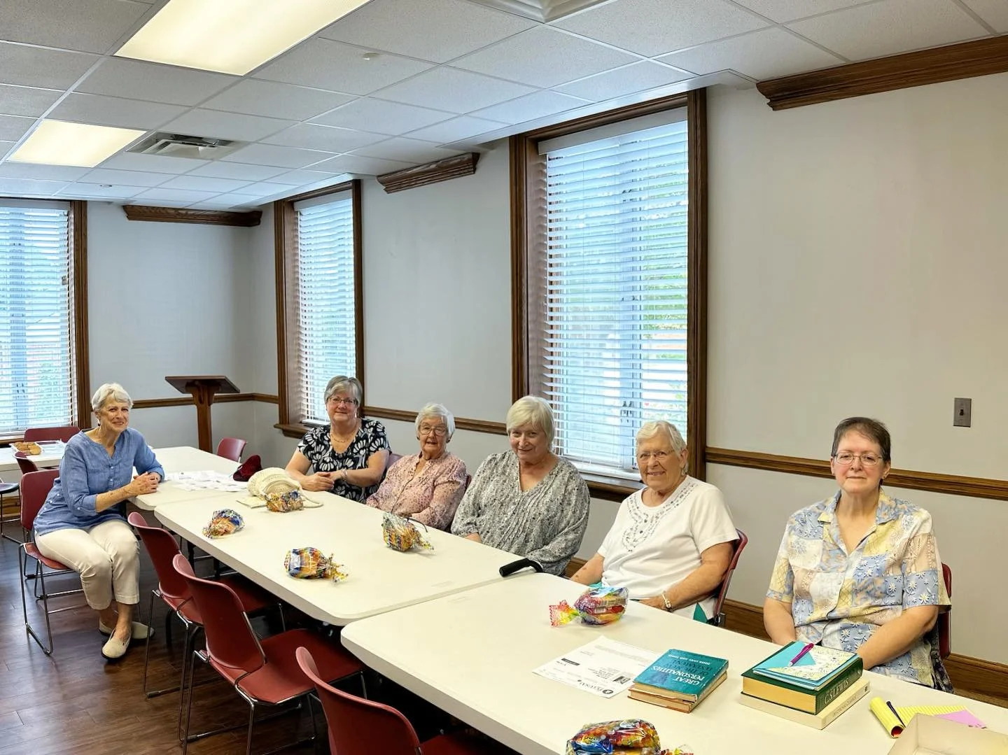 Seven elderly women sitting at a long meeting table in a well-lit room with large windows and wooden trim, with books, papers, and gift bags on the table.
