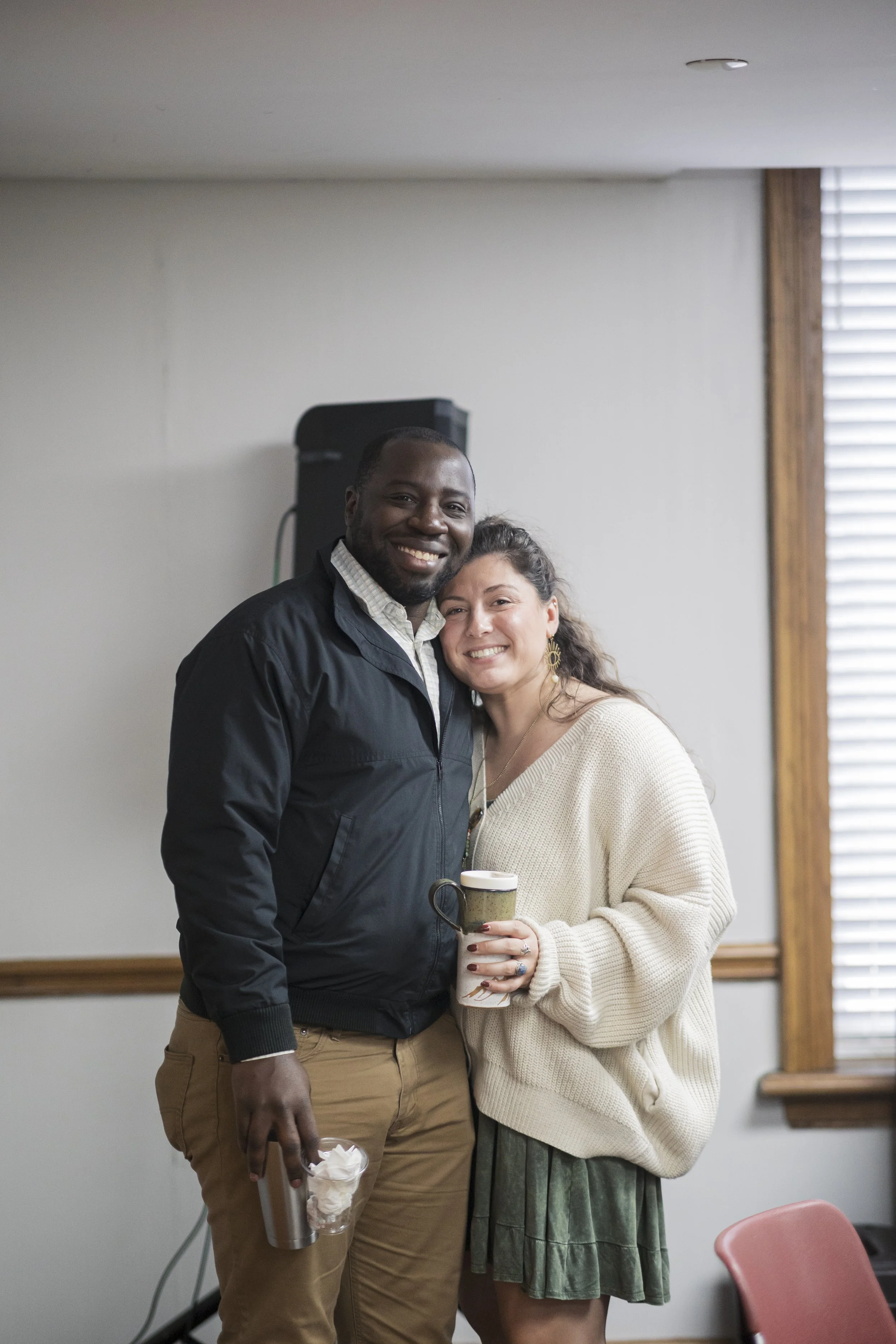 A smiling man and woman standing close together in a room, hug with the woman holding a mug, and the man holding a tumbler with whipped cream, with a window and a speaker in the background.