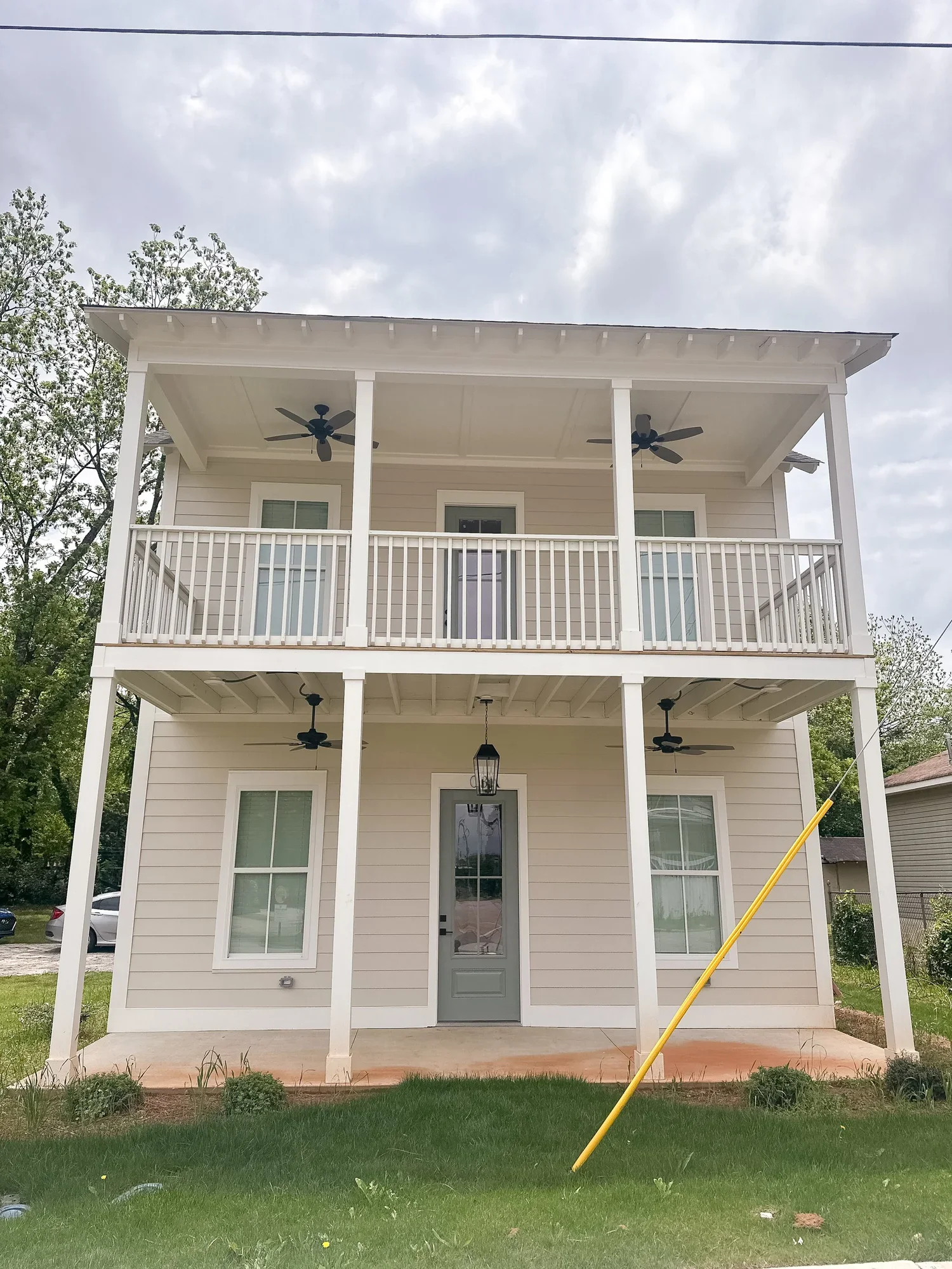 A three-story house with cream-colored siding, gray door, and two windows on the ground floor, decorated with black ceiling fans on each level, a hanging lantern above the front door, and a grassy yard with bushes and a yellow pole leaning against the house.