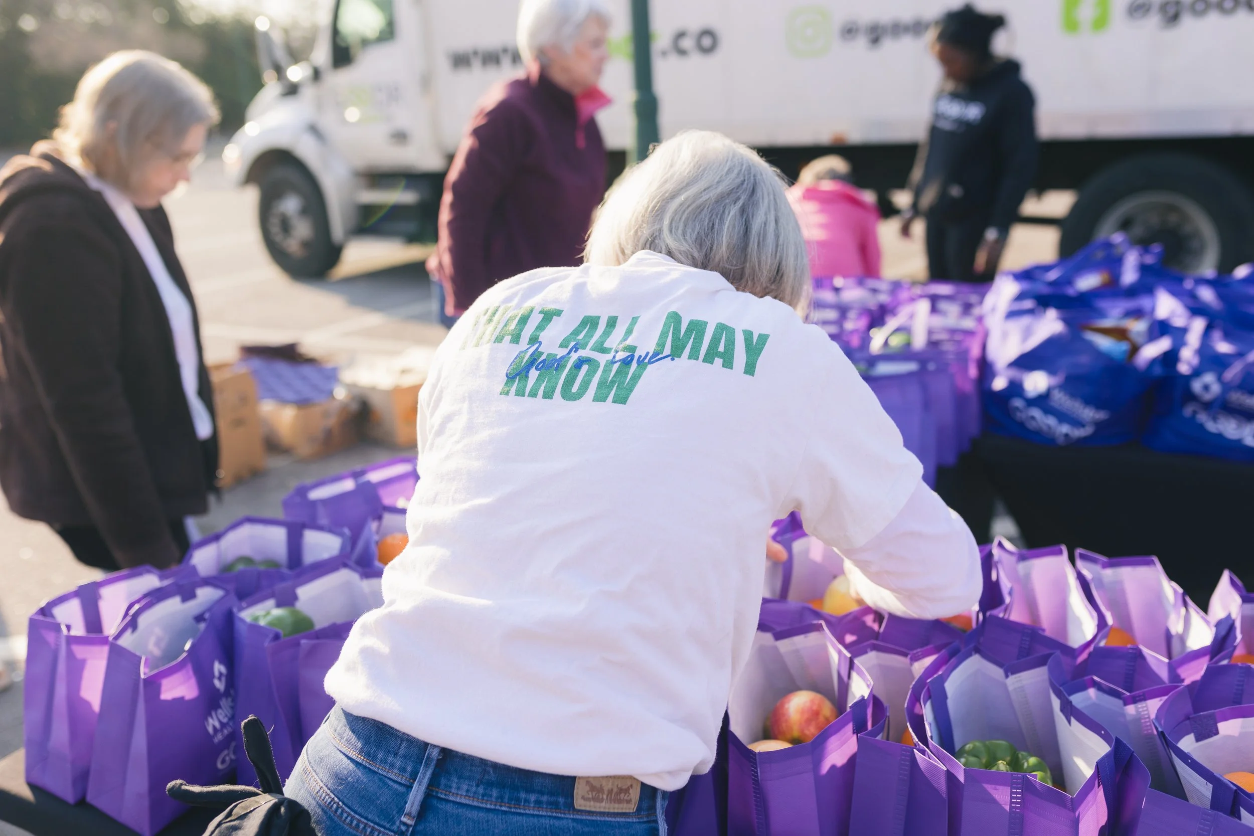 People distributing food or supplies at a community event, with purple bags filled with produce in the foreground, and a truck in the background.