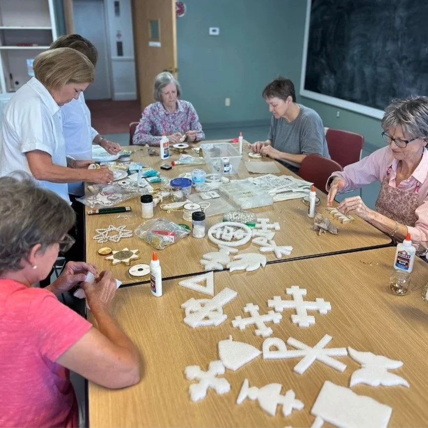 Group of women working on holiday-themed craft projects with white felt and decorative items at a table in a room with a chalkboard.