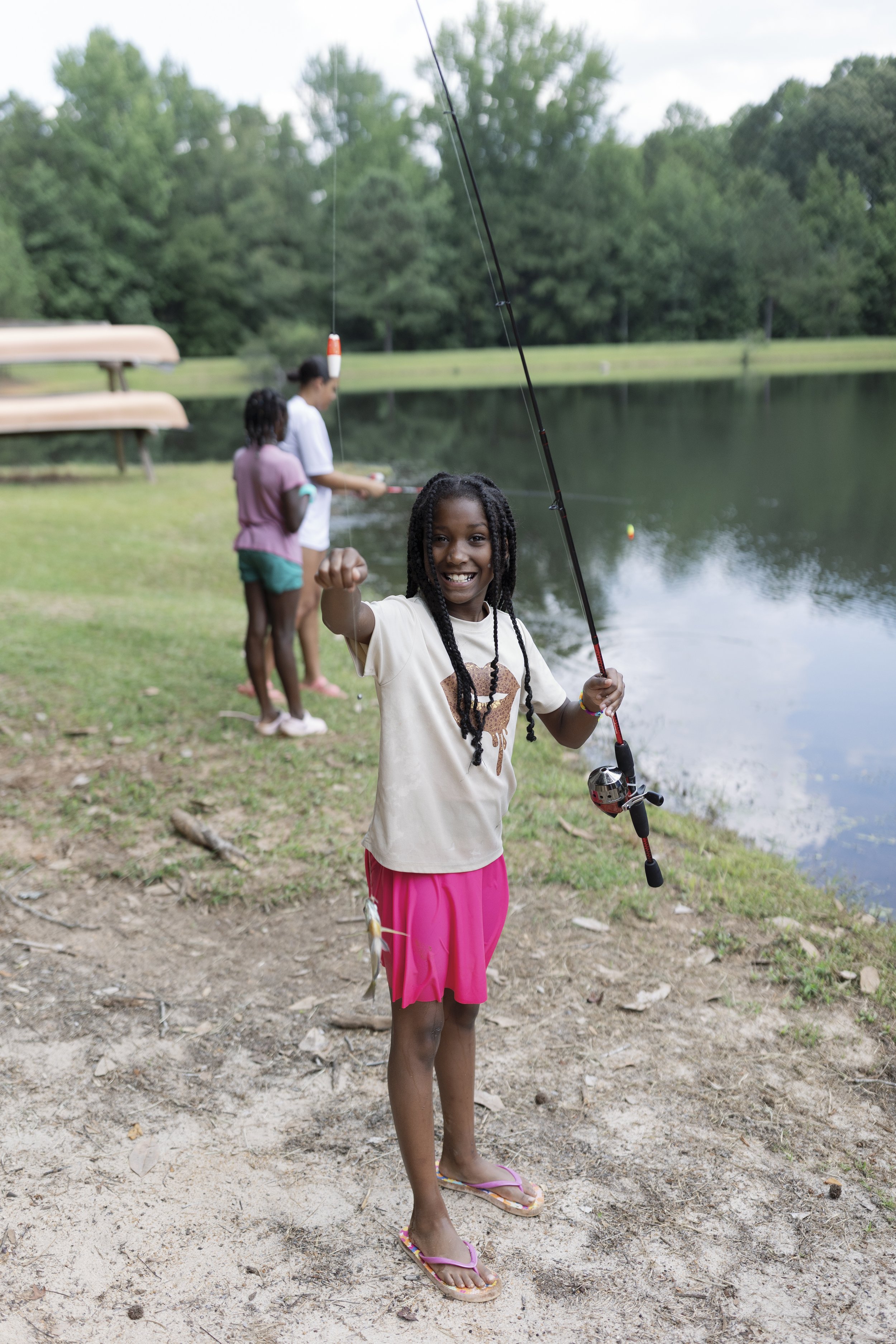 A group of children fishing by a lake, with a girl in the foreground holding a fishing rod and showing a fish she caught, smiling at the camera.