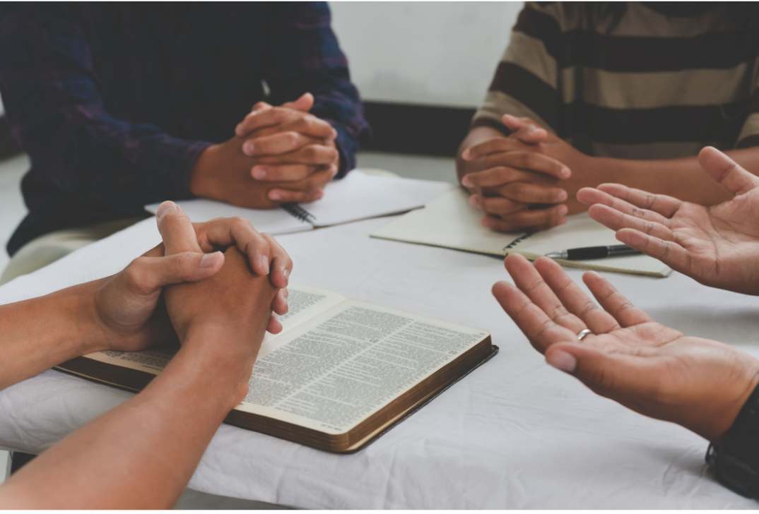 Four people sitting around a table engaged in prayer, with an open Bible, notebooks, and a pen visible.
