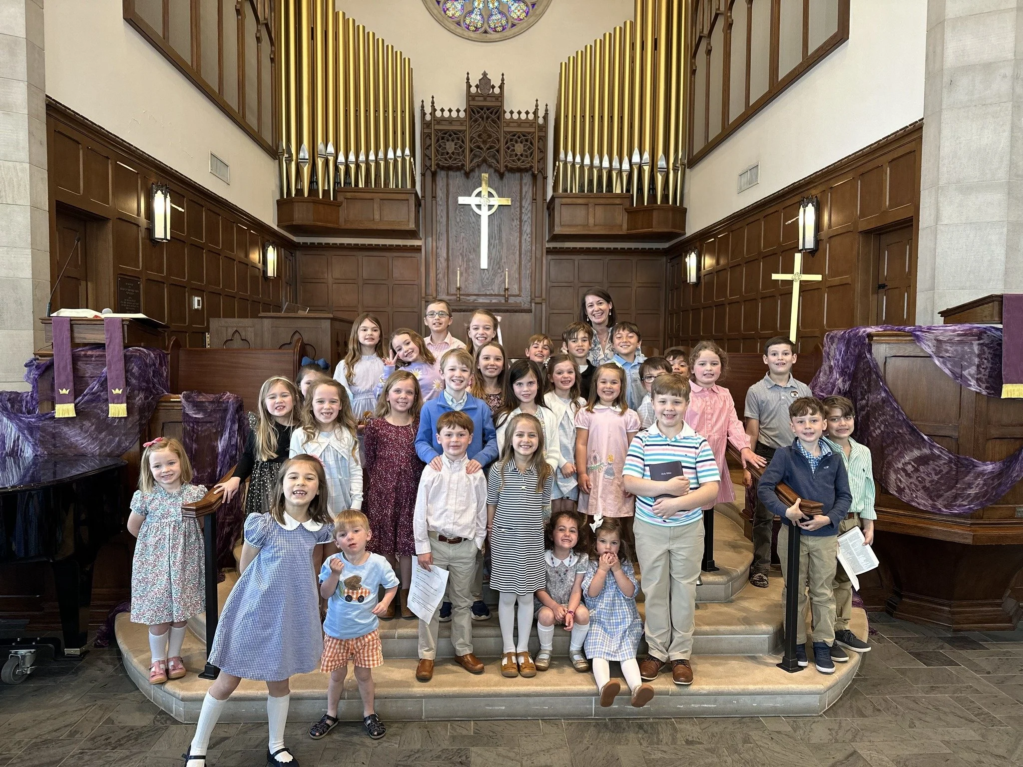 A group of children and a woman standing on the steps of an altar inside a church with wooden paneling, gold organ pipes, a cross, and a stained glass window.