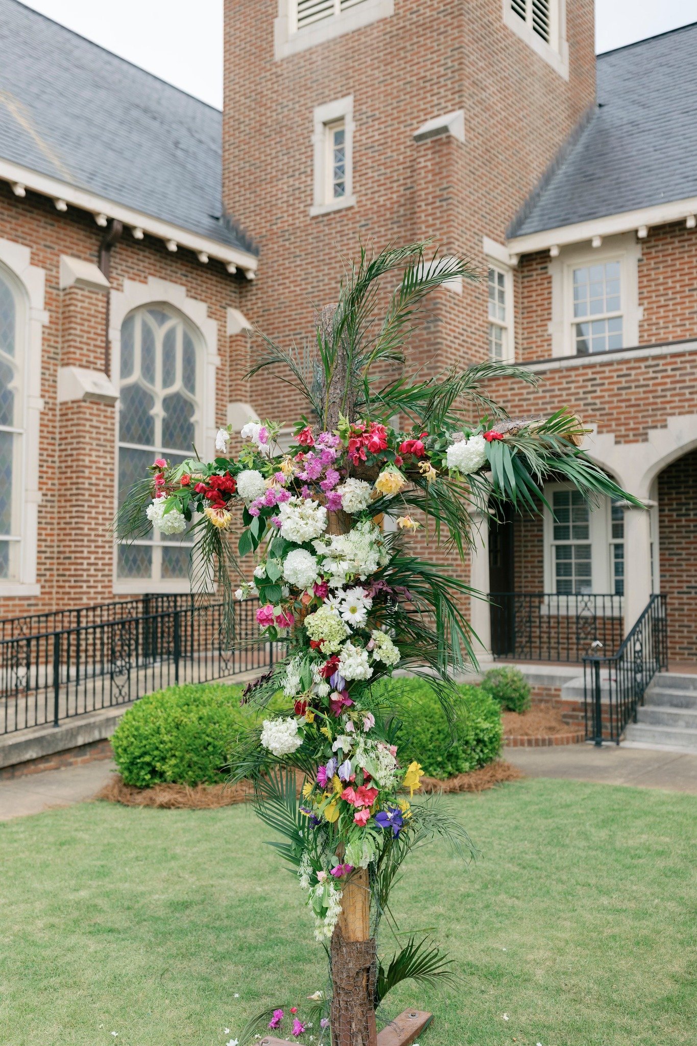 A decorative floral arrangement shaped like a cross with a variety of colorful flowers and green foliage, placed outside a brick church or building.