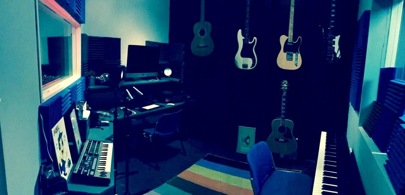 Music studio with guitars hanging on the wall, a keyboard, computer, and sound equipment on desks.